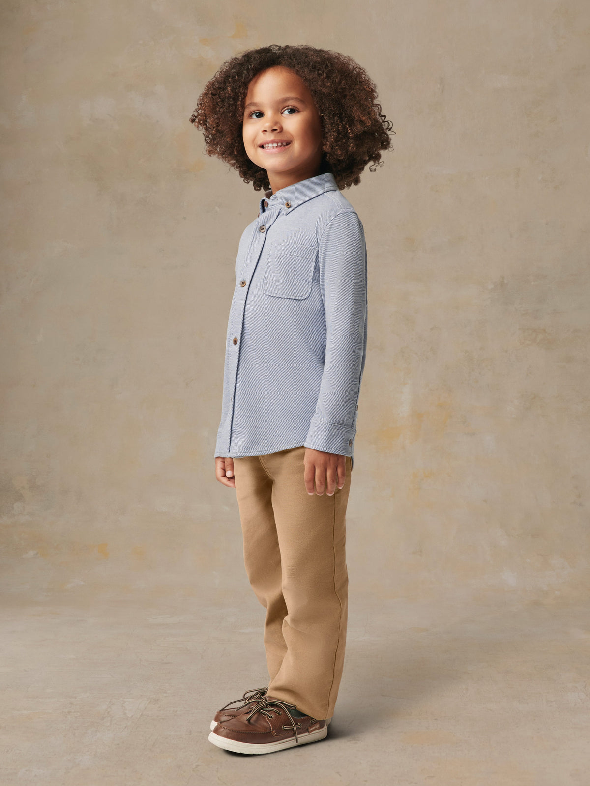 A child wearing a blue button up shirt and beige pants, image 5 of 6