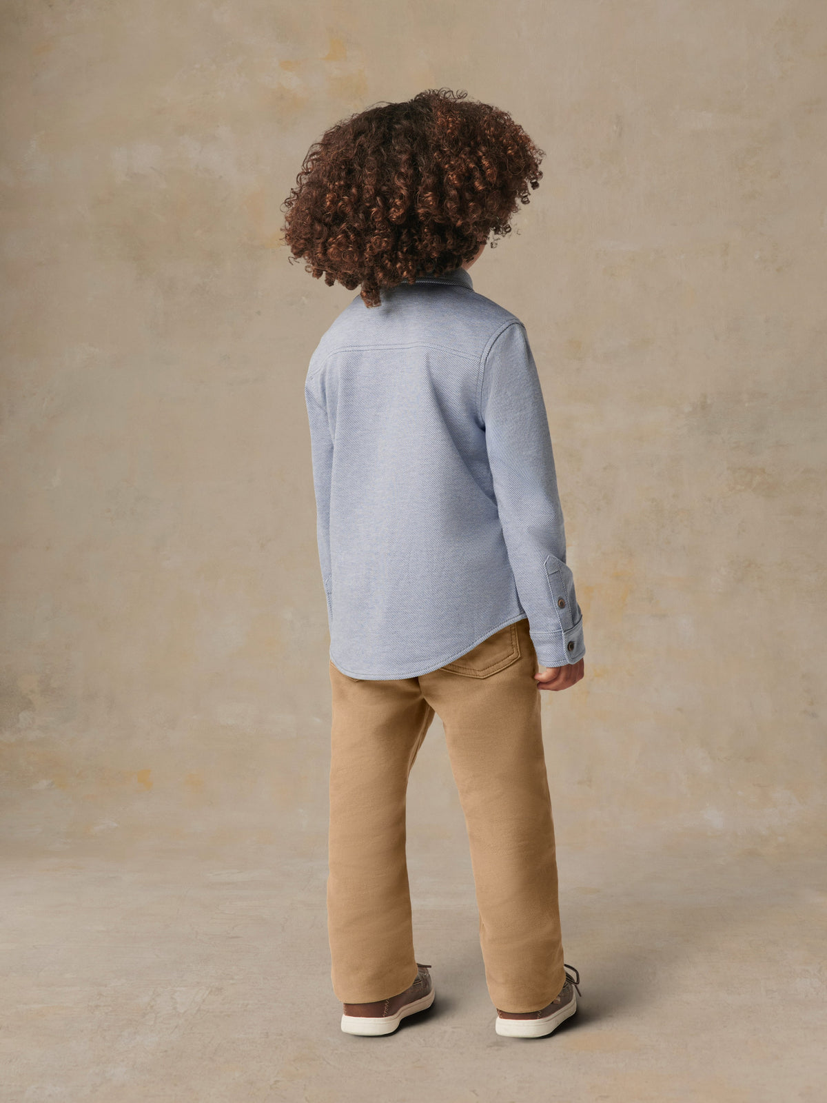 A child viewed from behind wearing a blue button up shirt and beige pants, image 3 of 6