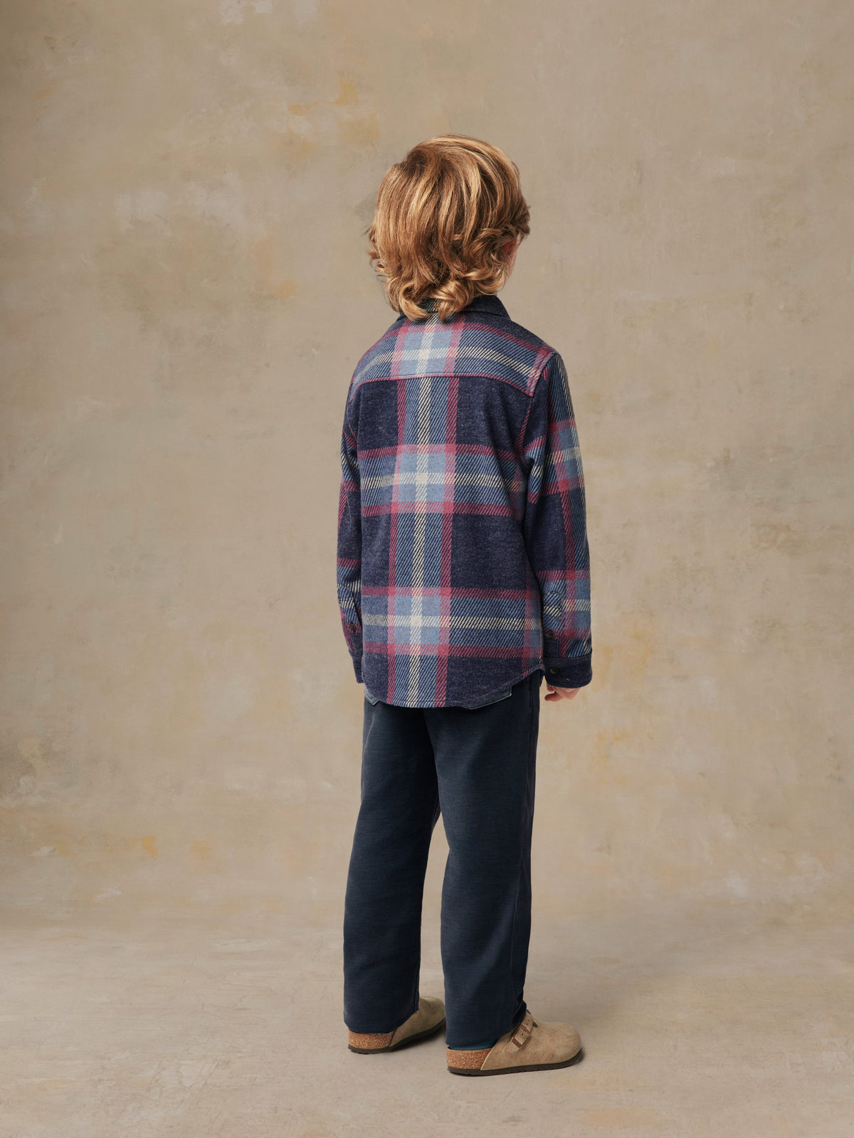 A child viewed from behind wearing a plaid shirt and black pants, image 3 of 6