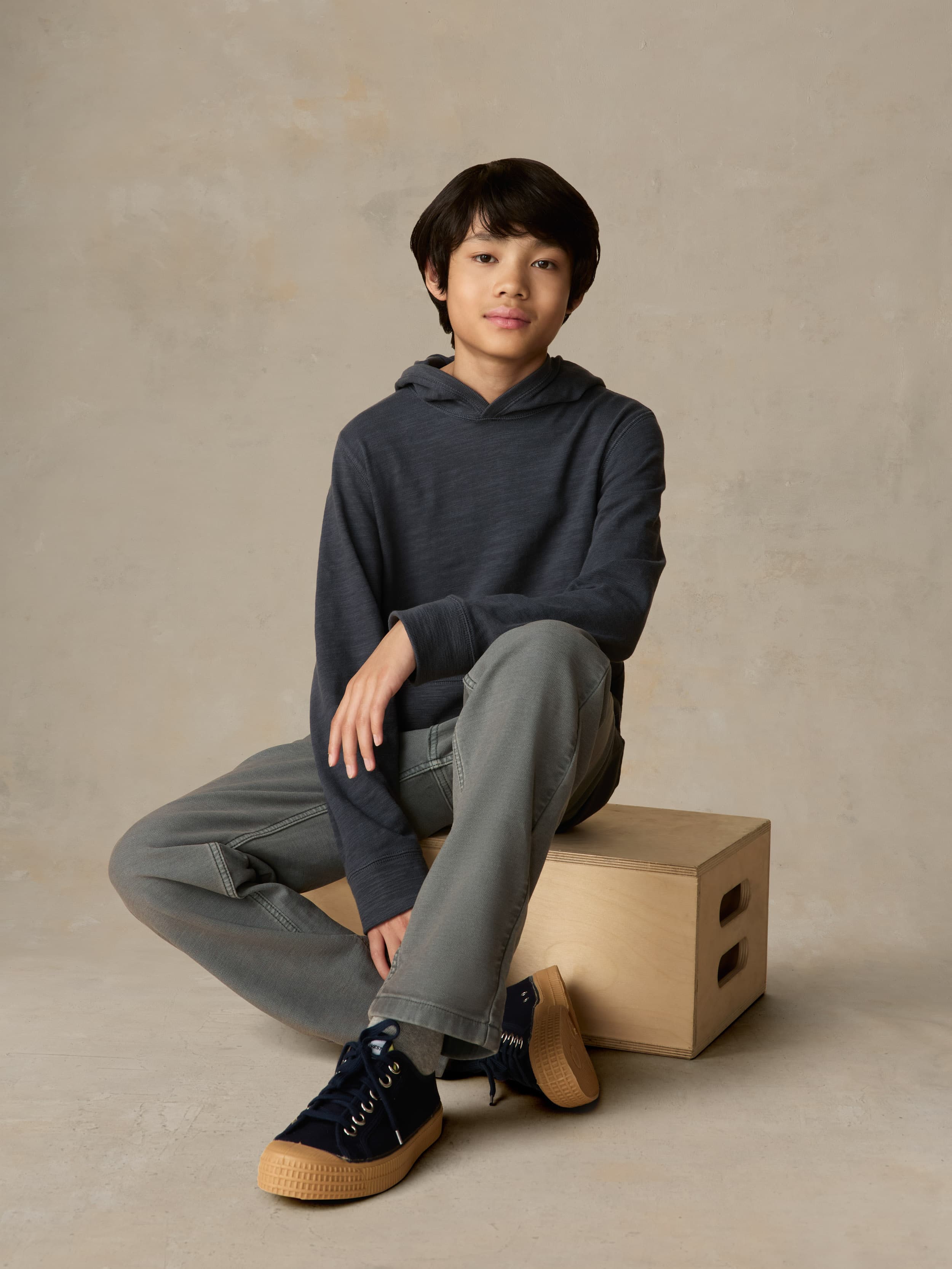 Child sitting on a wooden box wearing a blue hoodie with grey pants against a beige background