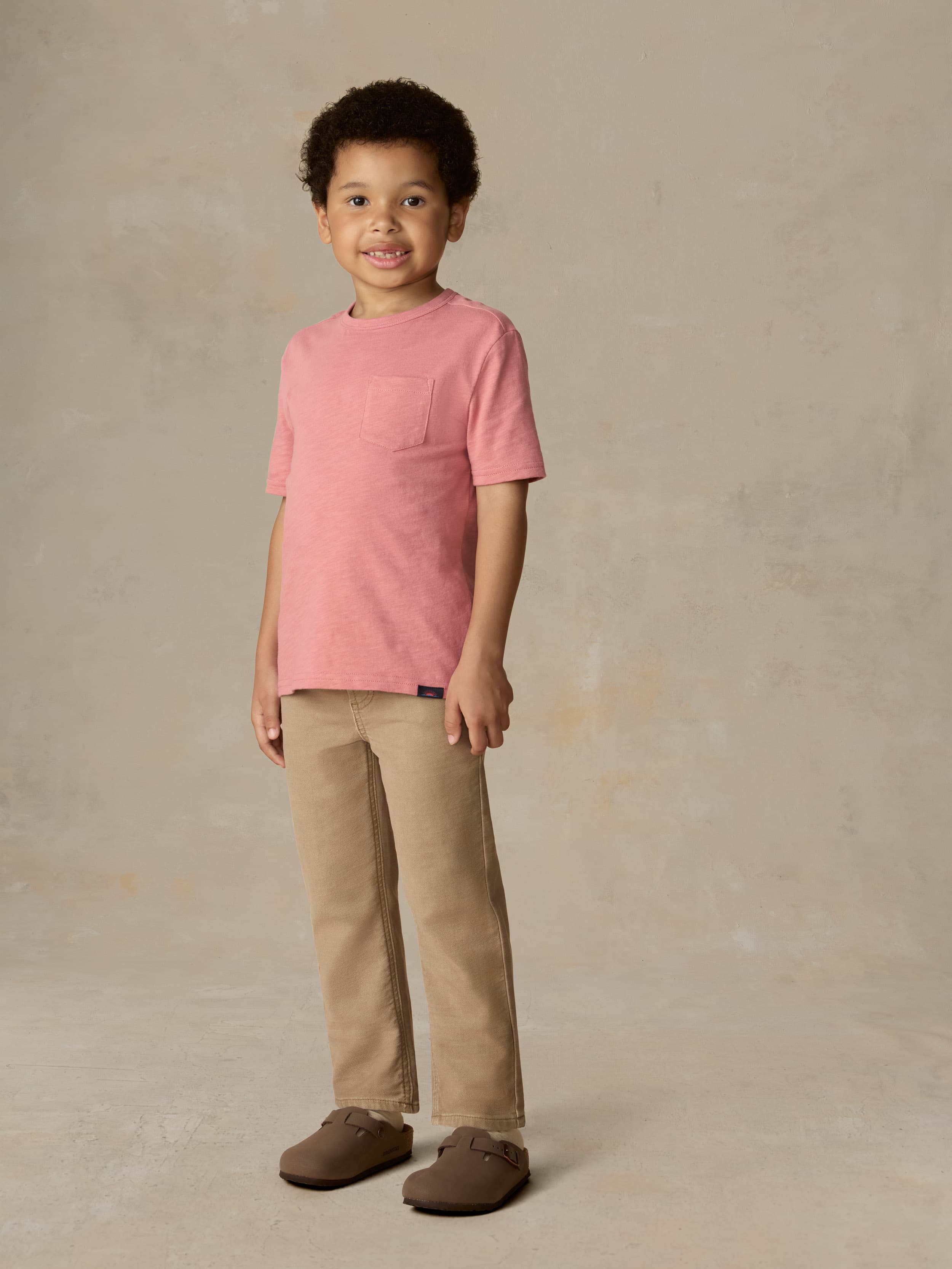 Child wearing a red t-shirt and beige pants against a beige background