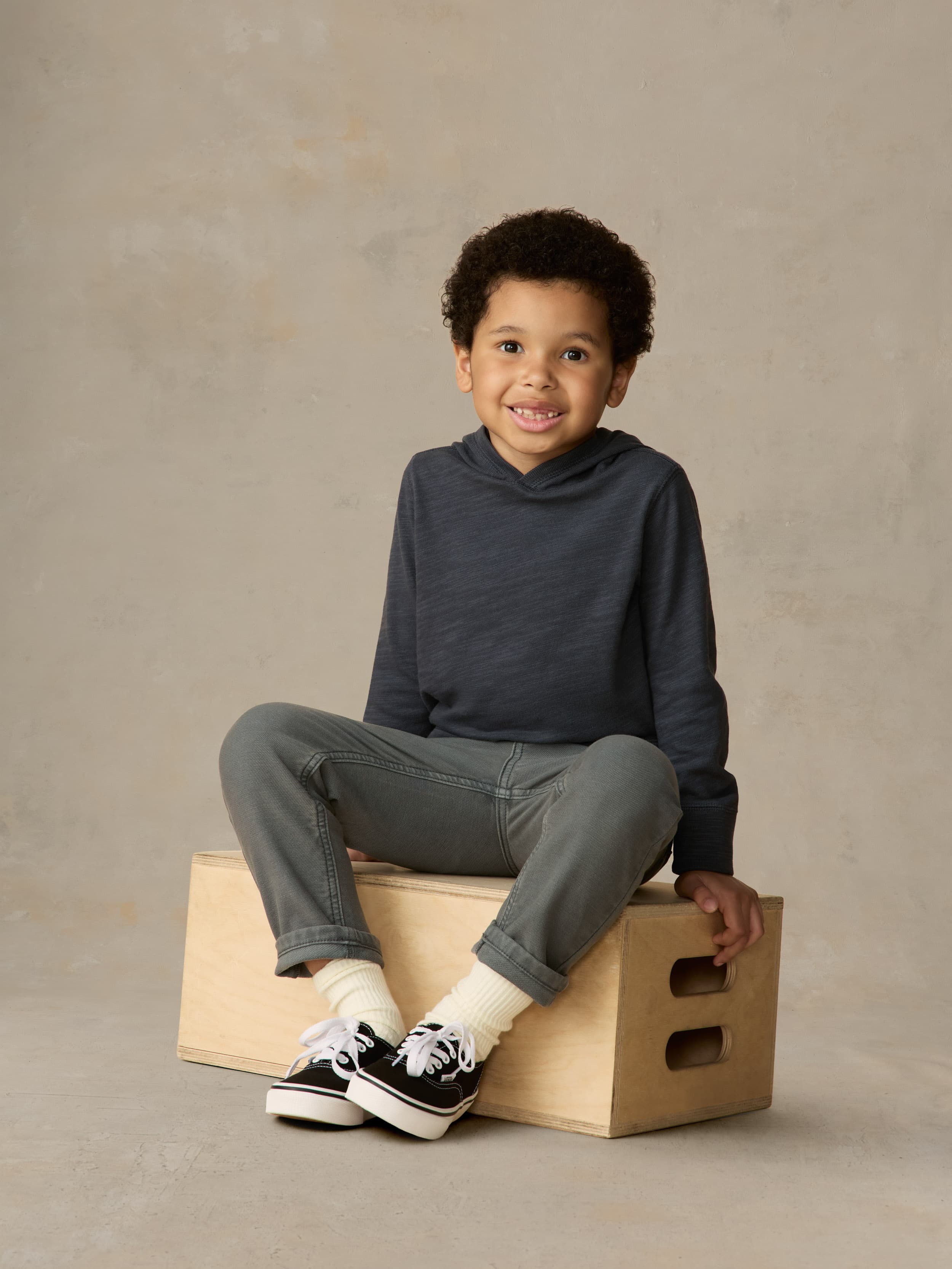 Child sitting on a wooden box wearing blue hoodie and grey pants against a beige background