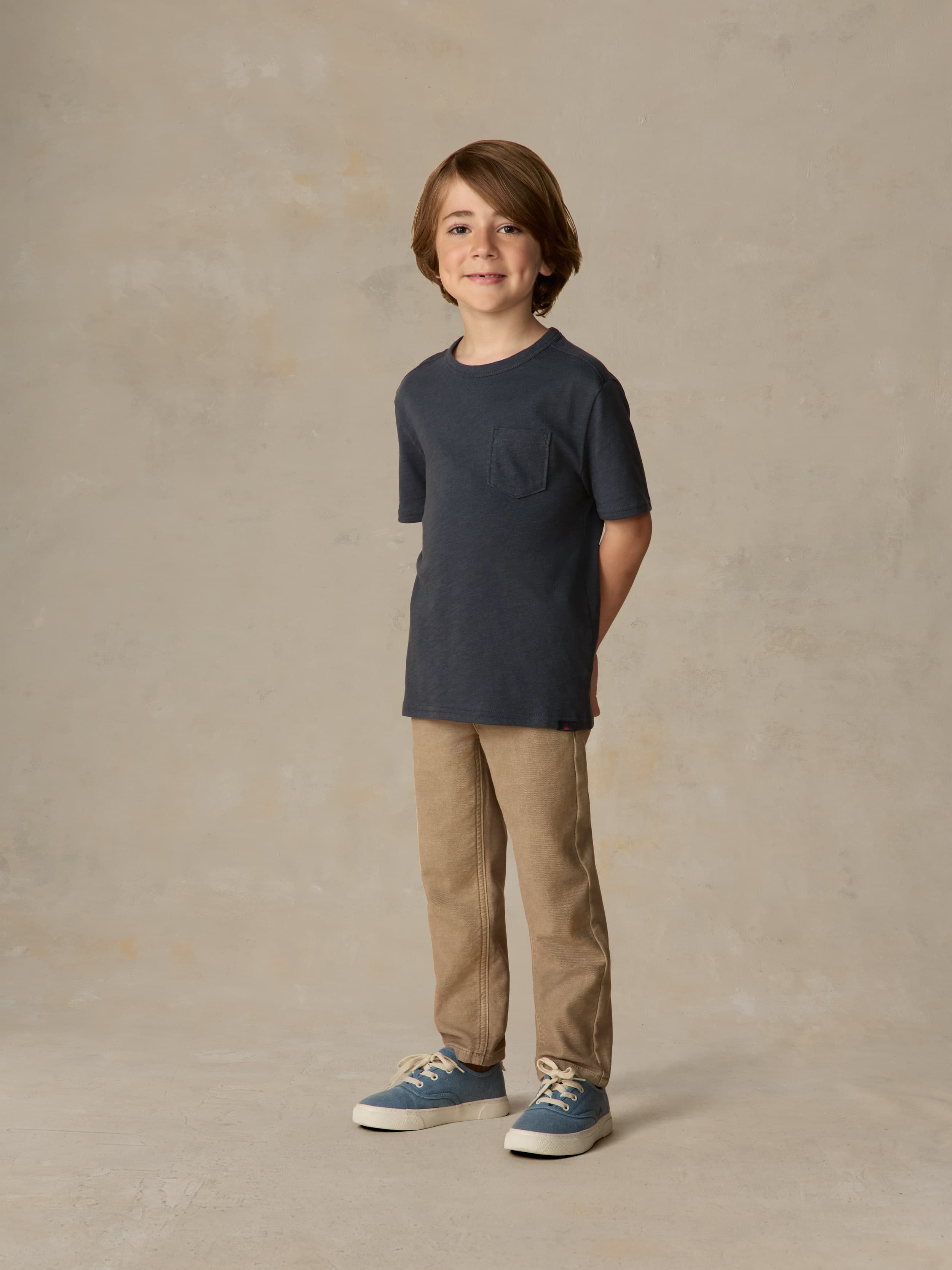 Child wearing a blue pocket tee and beige pants against a beige background