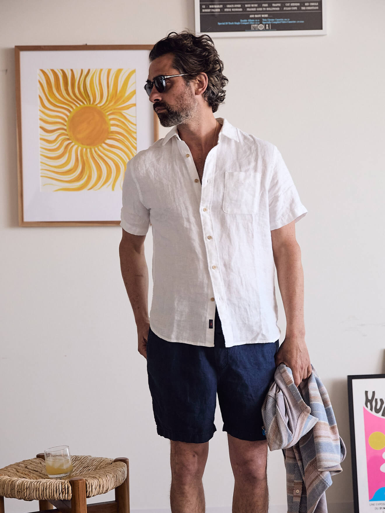 Male model wearing a white linen shirt and blue linen shorts, holding a legend sweater shirt in his hand
