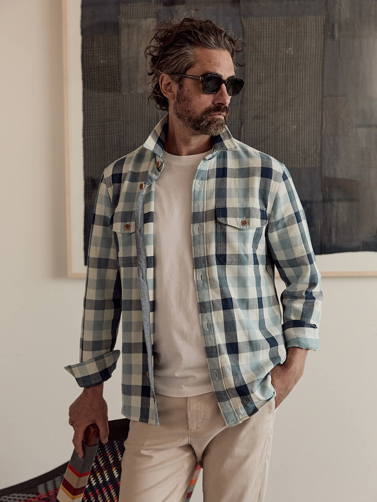 A male model wearing a plaid work shirt over a white tee with sunglasses and white chinos
