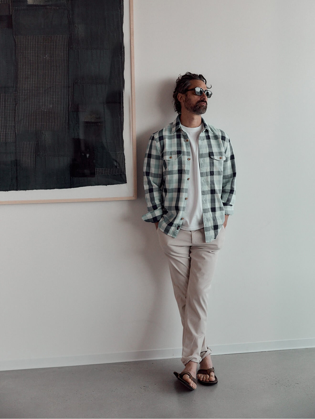 A male model wearing a plaid work shirt over a white tee, with white chino pants and sandals