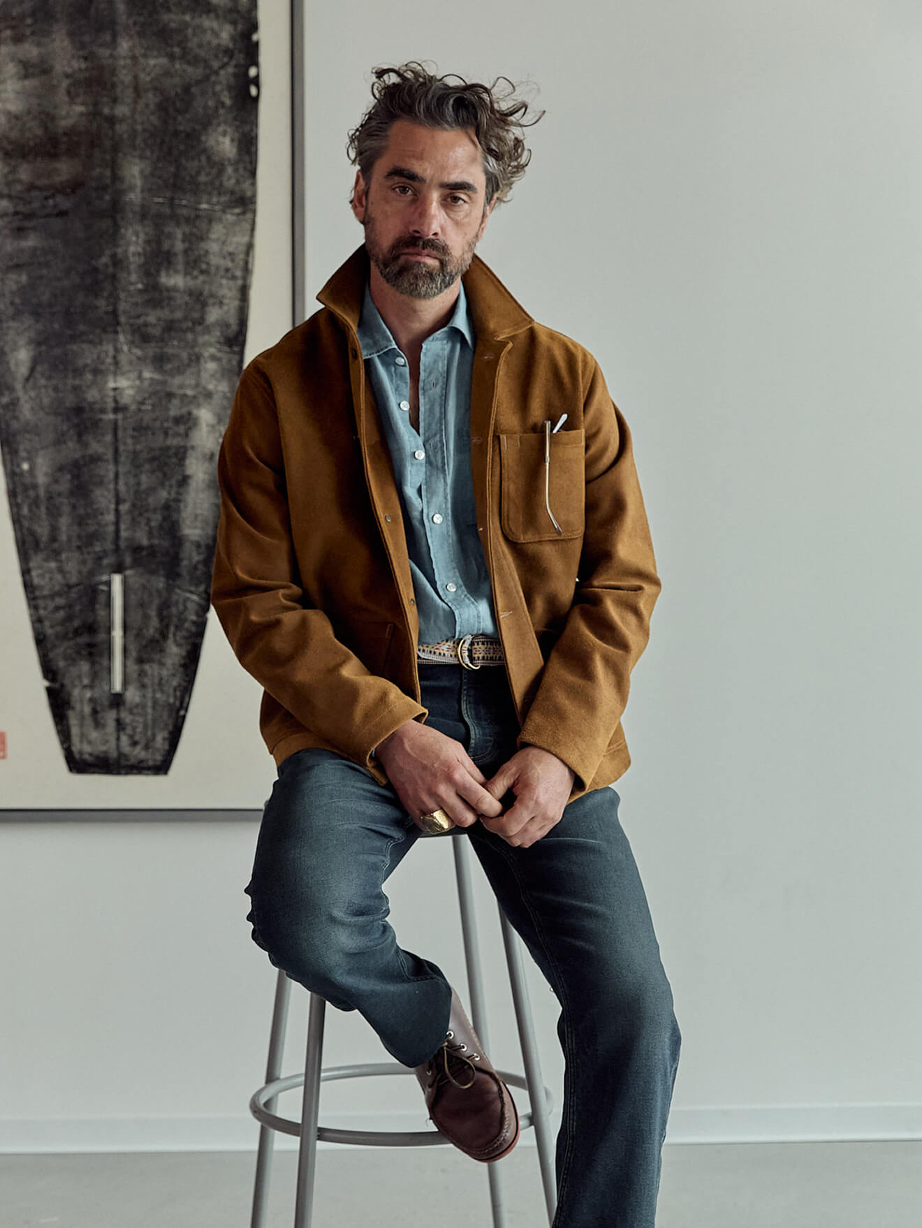 A seated male model wearing a suede studio jacket over a chambray shirt, paired with indigo stretch terry pants and quoddy boat shoes