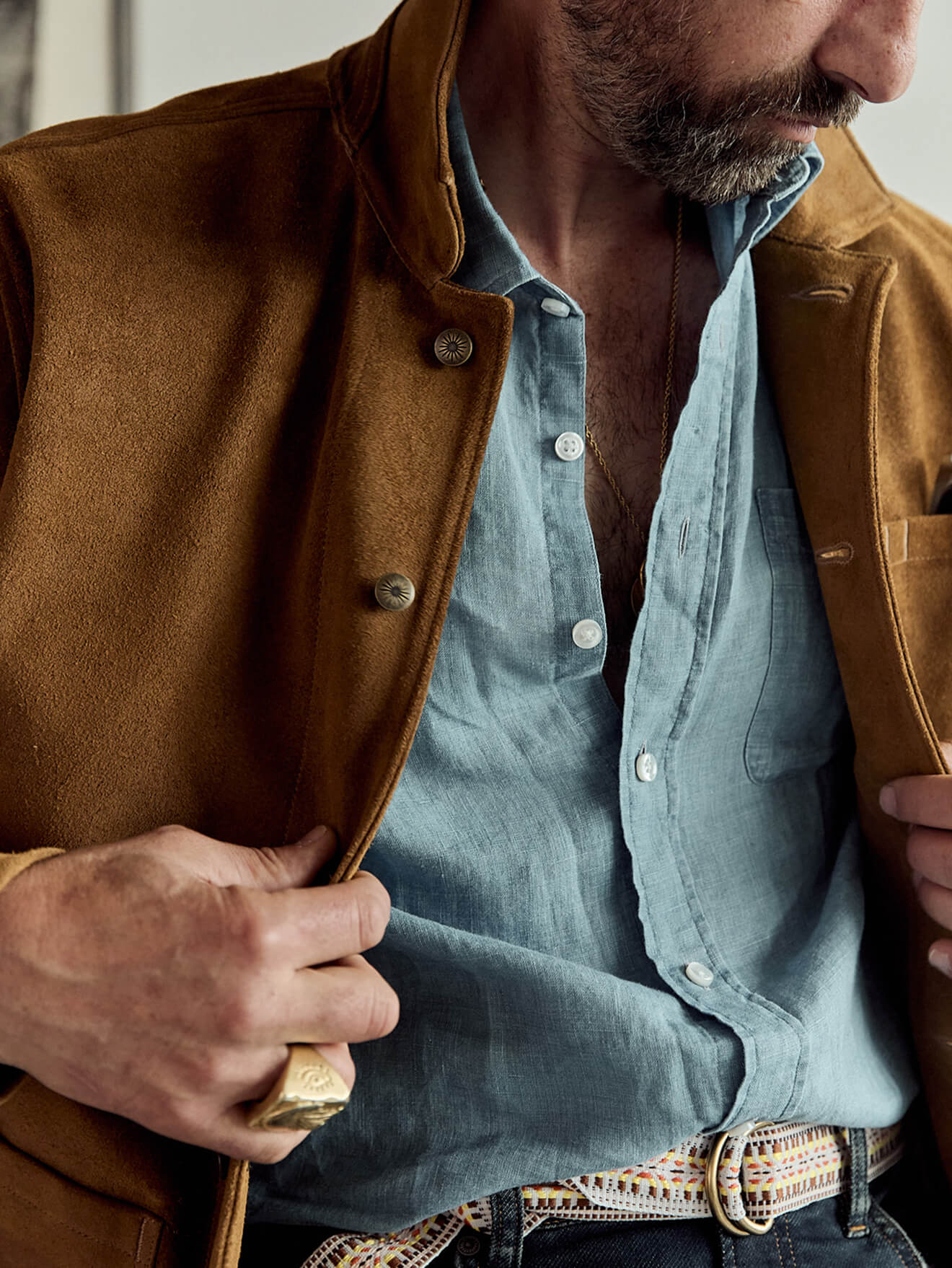 A seated male model wearing a suede studio jacket over a chambray shirt, paired with indigo stretch terry pants
