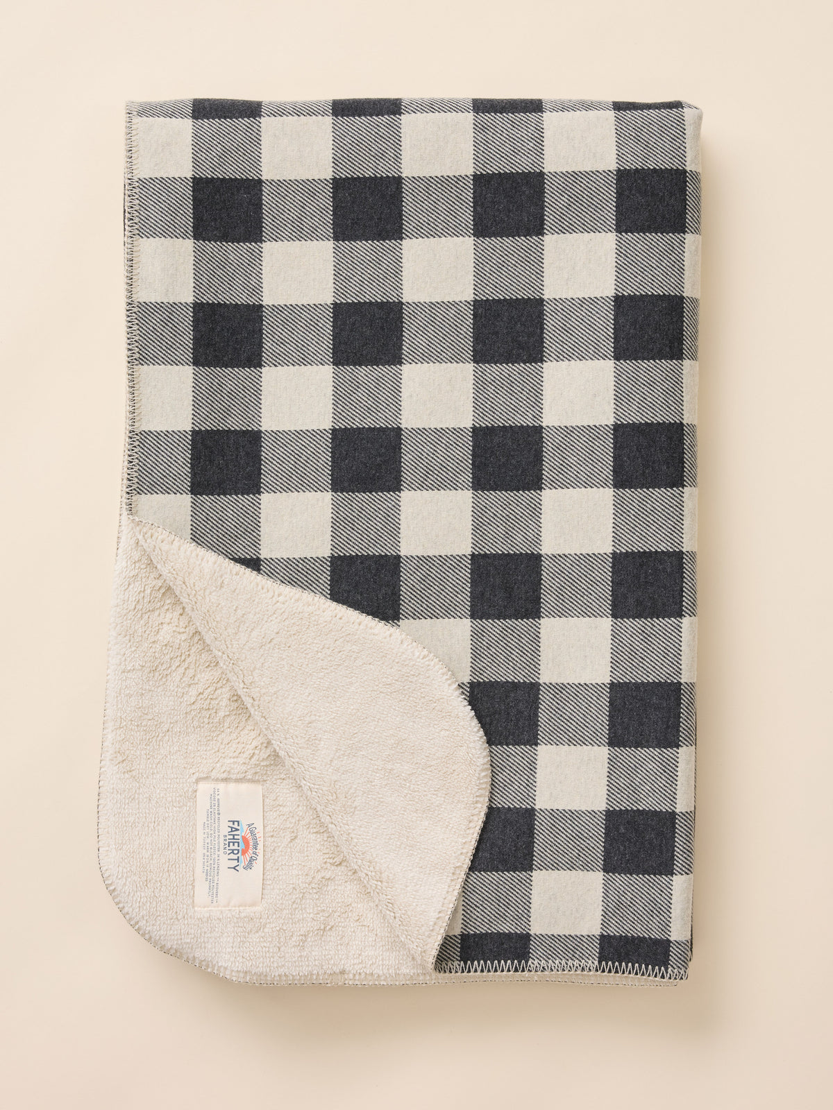 A folded checkered blanket with a black and off-white buffalo plaid pattern, showing a soft off-white fleece lining on the reverse side, photographed on a solid light beige background., image 1 of 3