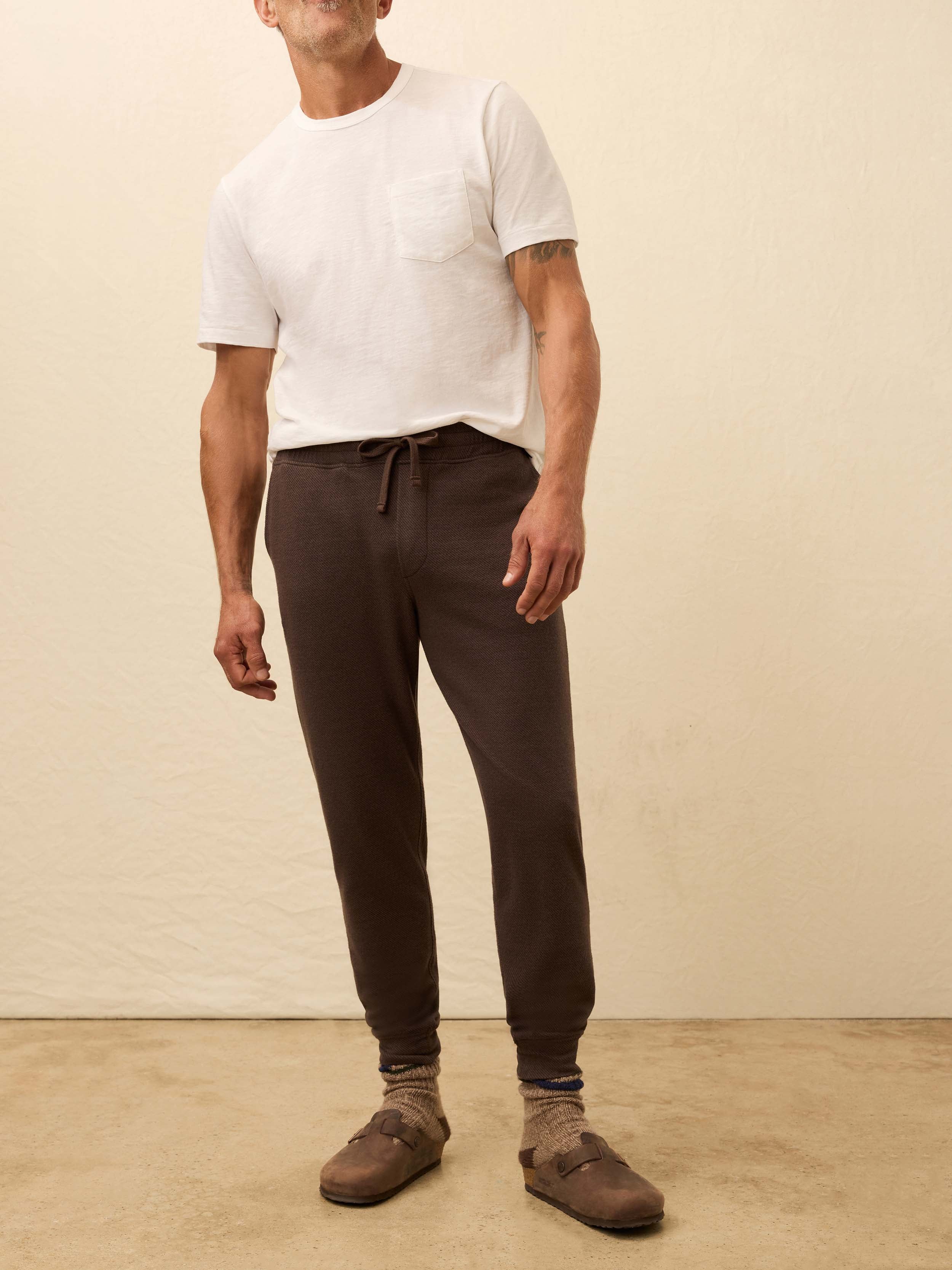 Man wearing a white t-shirt and brown sweatpants against a beige background