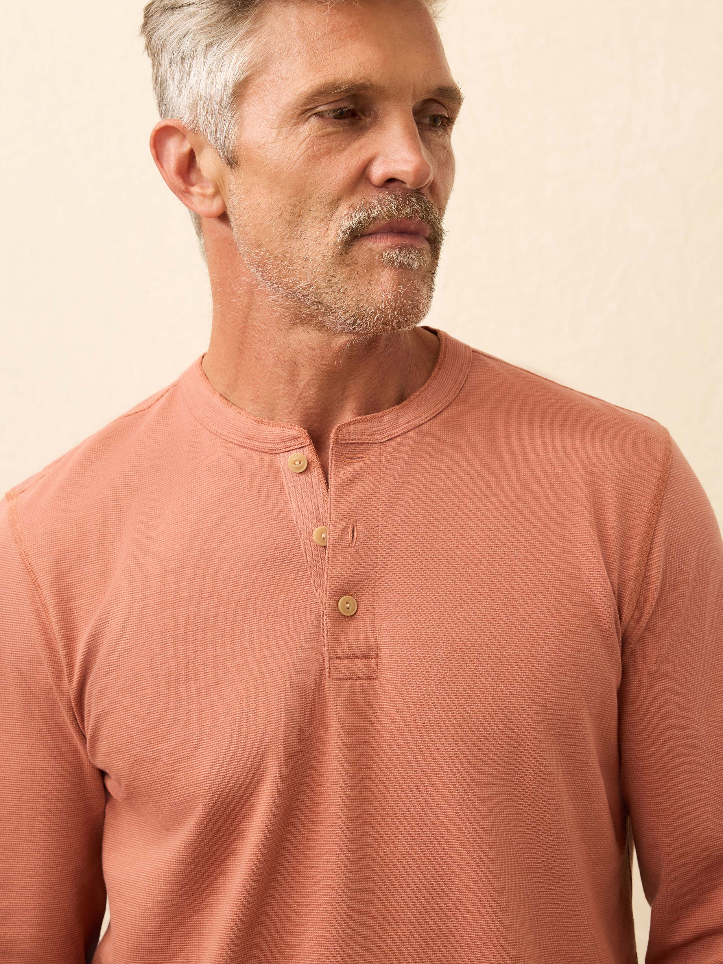 Man wearing a salmon color long sleeve henley shirt against a beige background