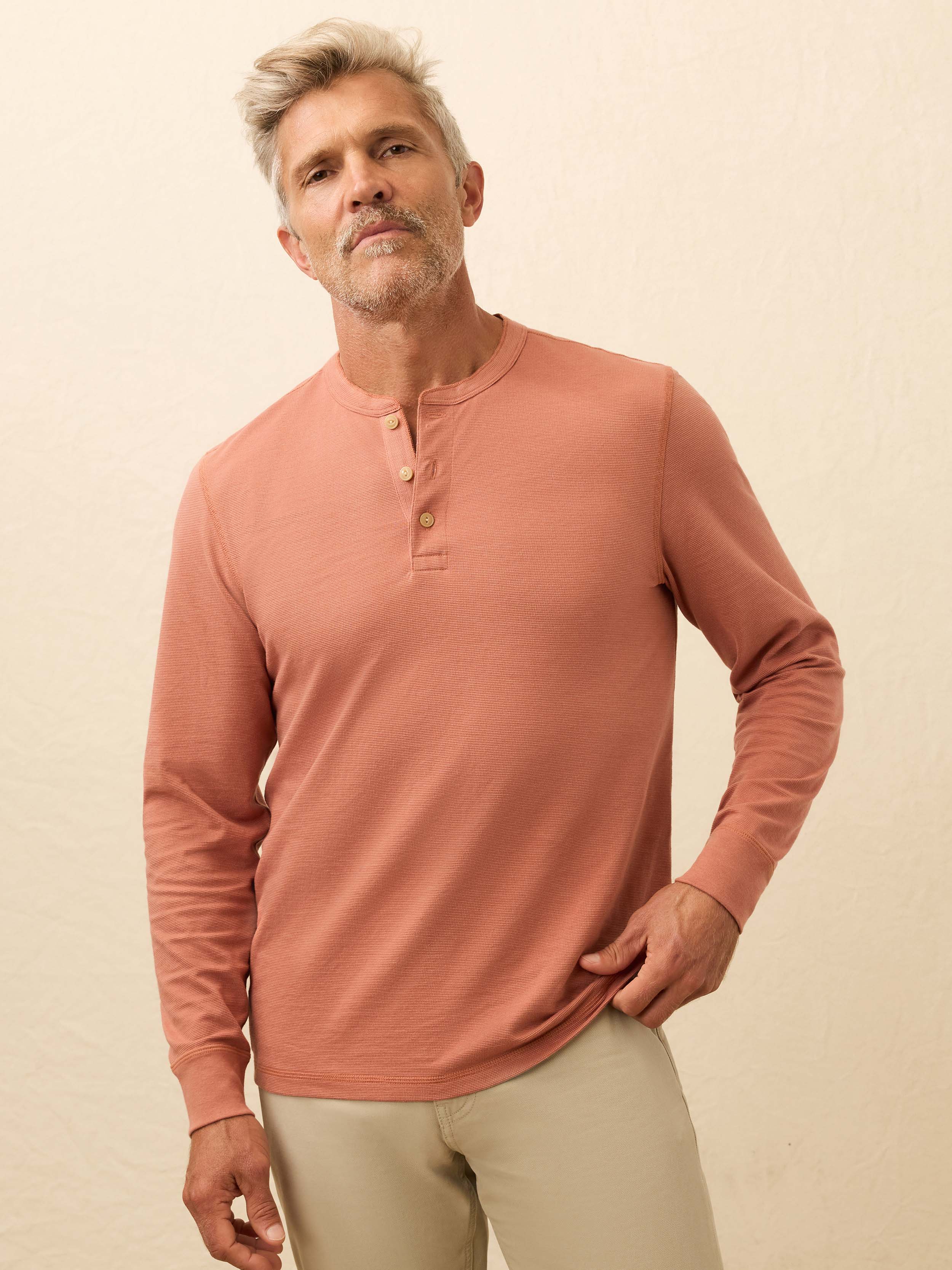 Man wearing a salmon color long sleeve henley shirt against a beige background