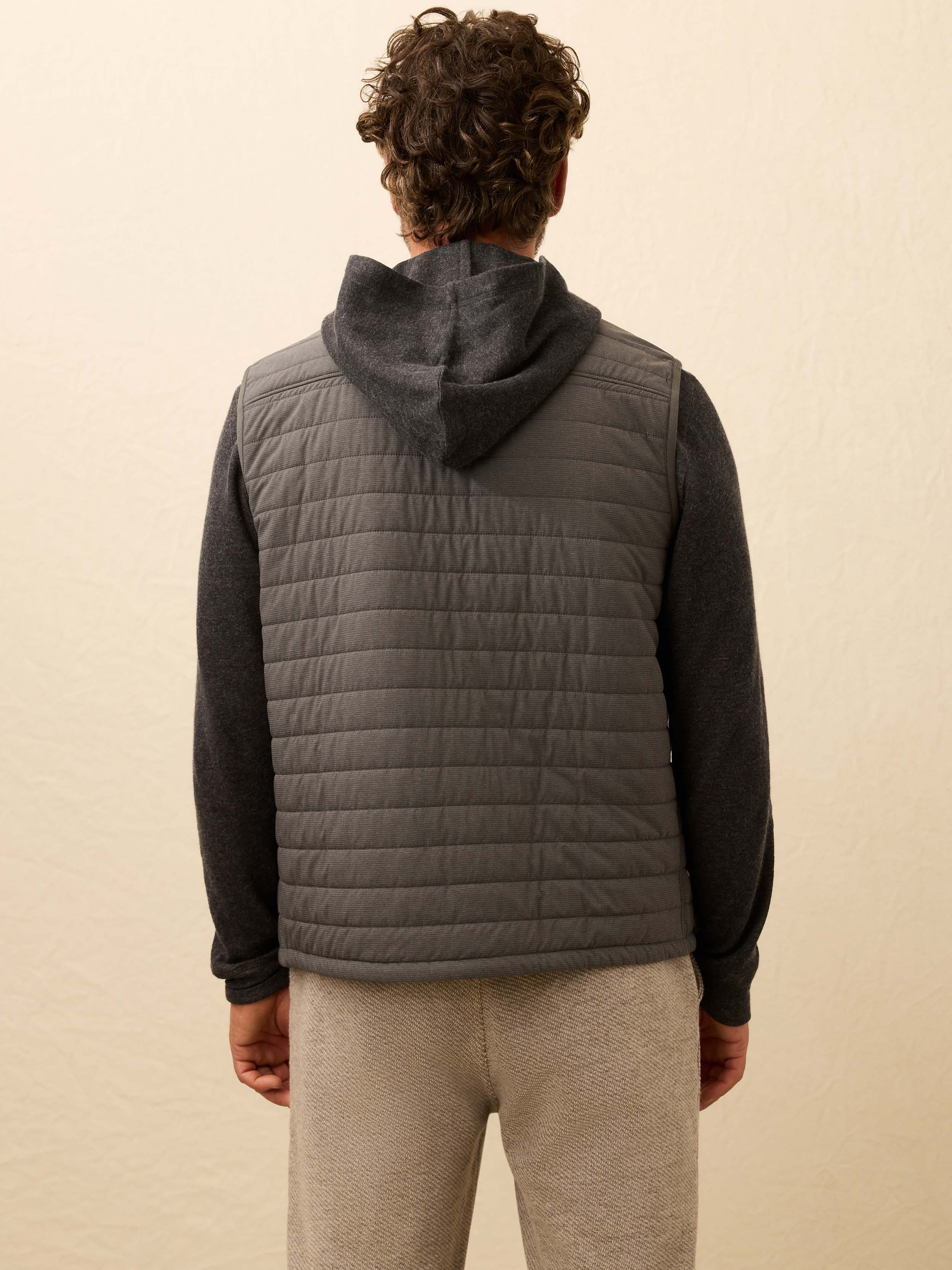Man wearing a grey quilted vest over a dark grey hoodie against a beige background