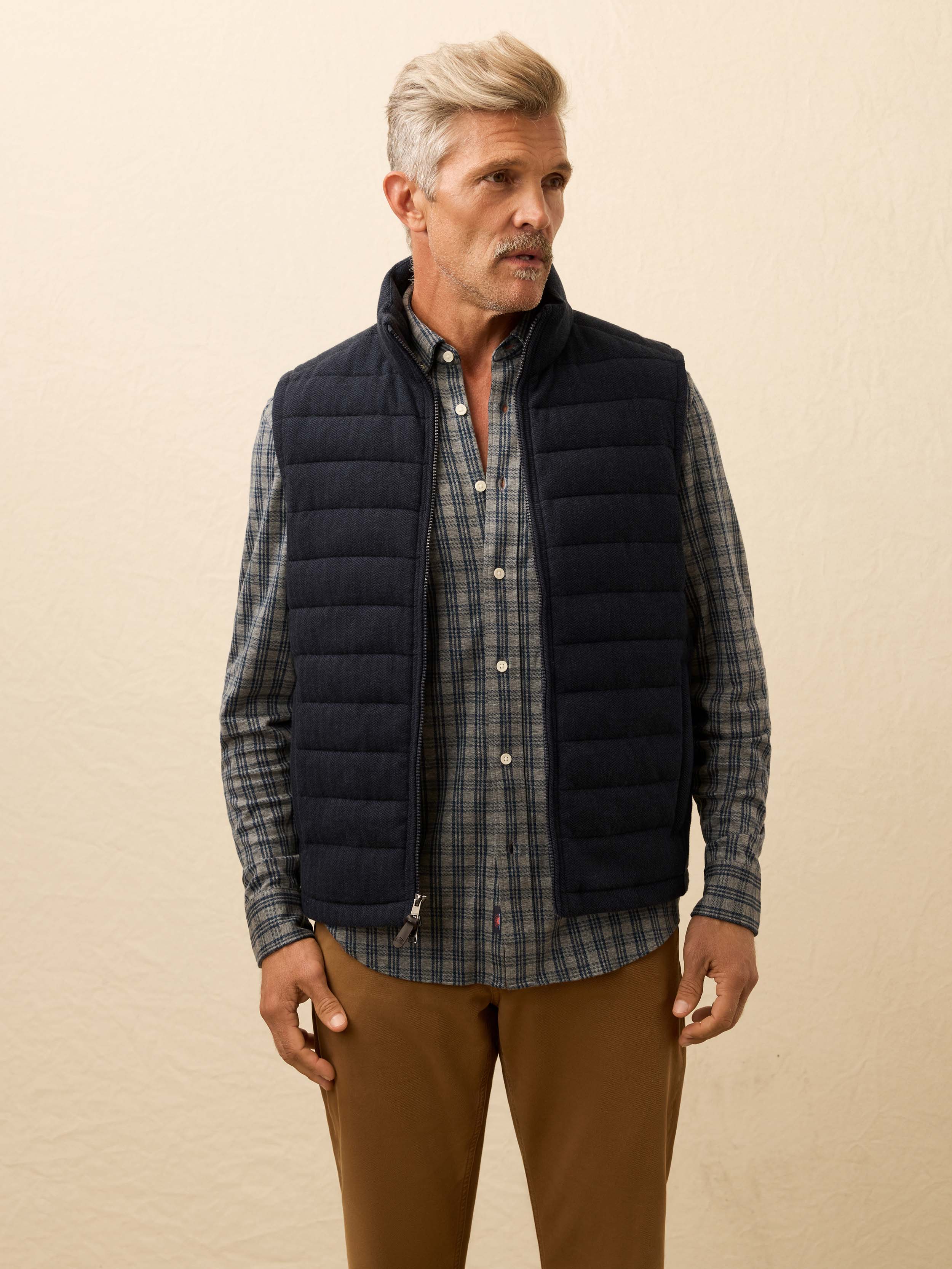 Man is wearing a navy quilted vest layered over a grey plaid shirt against a beige background