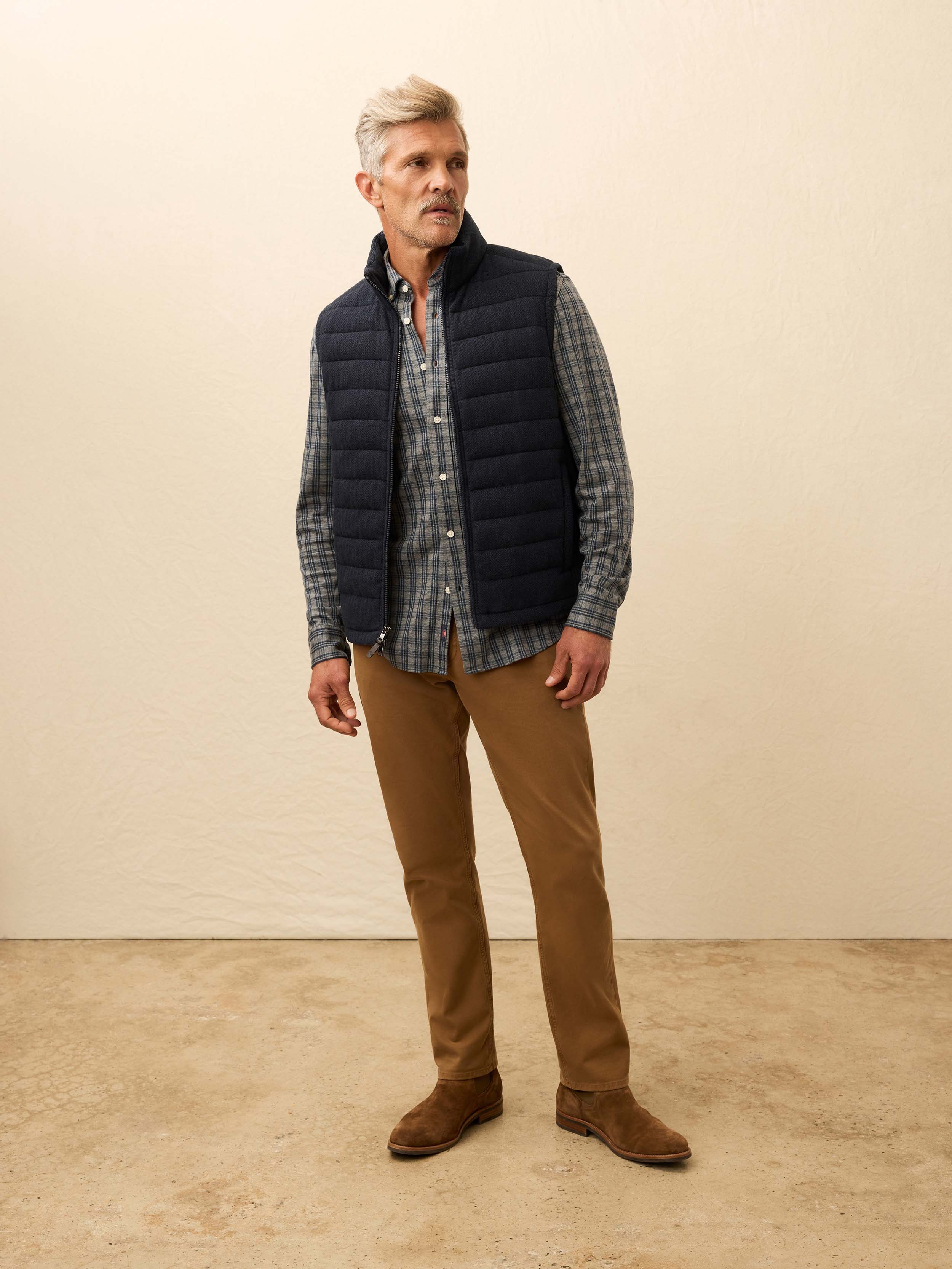 Man is wearing a navy quilted vest layered over a grey plaid shirt against a beige background