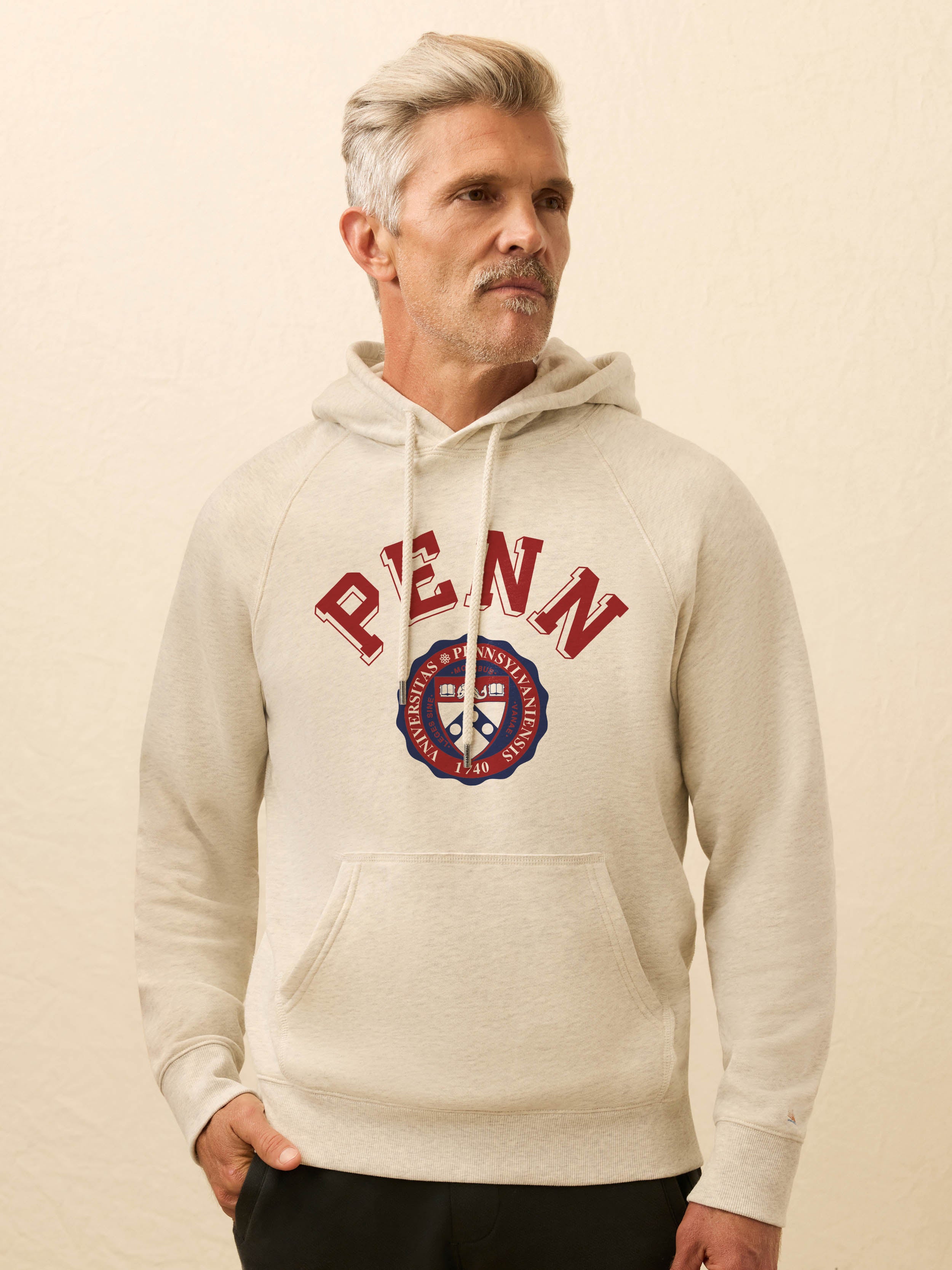 Man is wearing a heather grey hoodie with University of Pennsylvania graphic on the front against a beige background