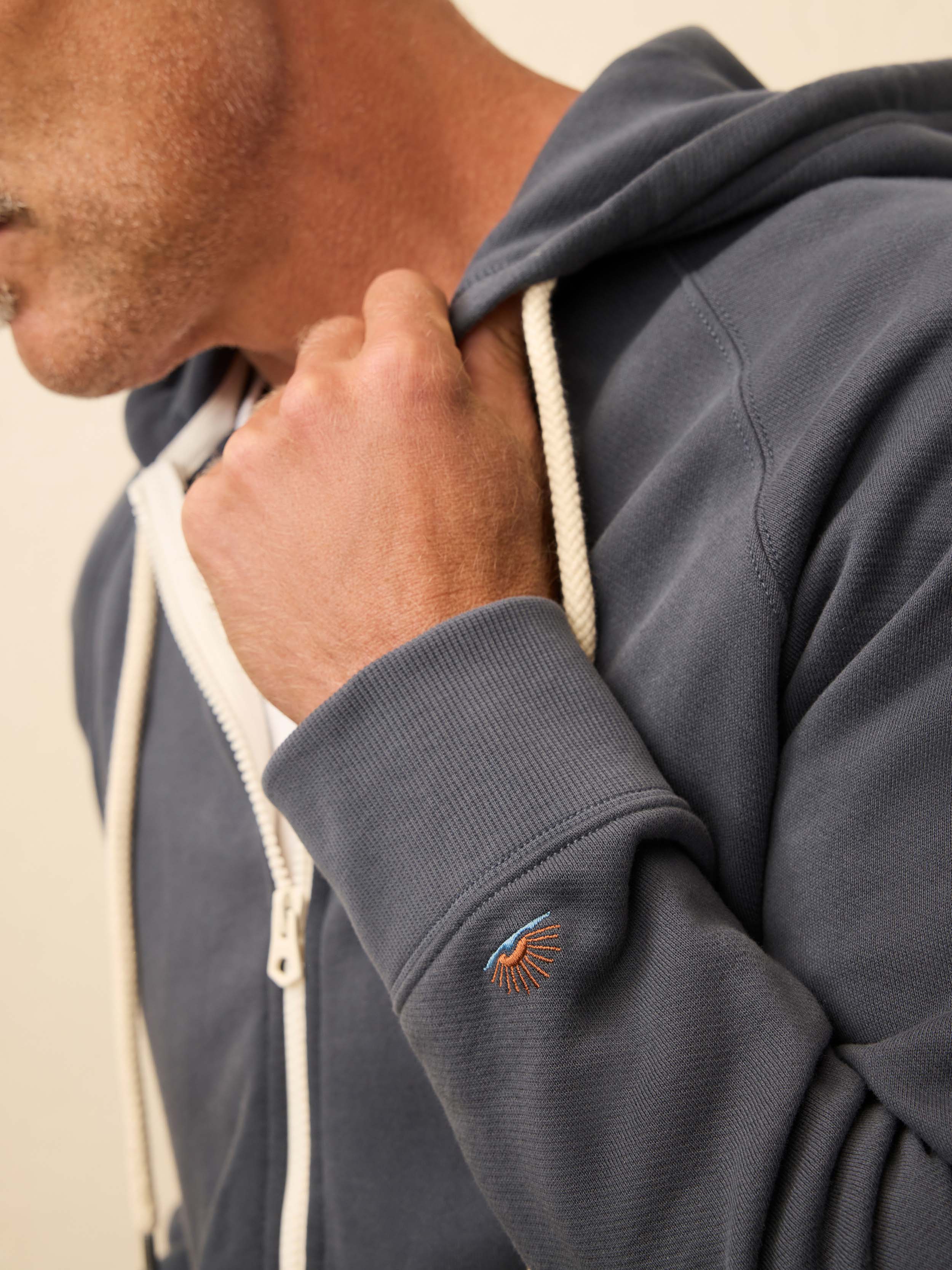 Man is wearing a navy zip hoodie with Faherty branded logo on the wrist against a beige background