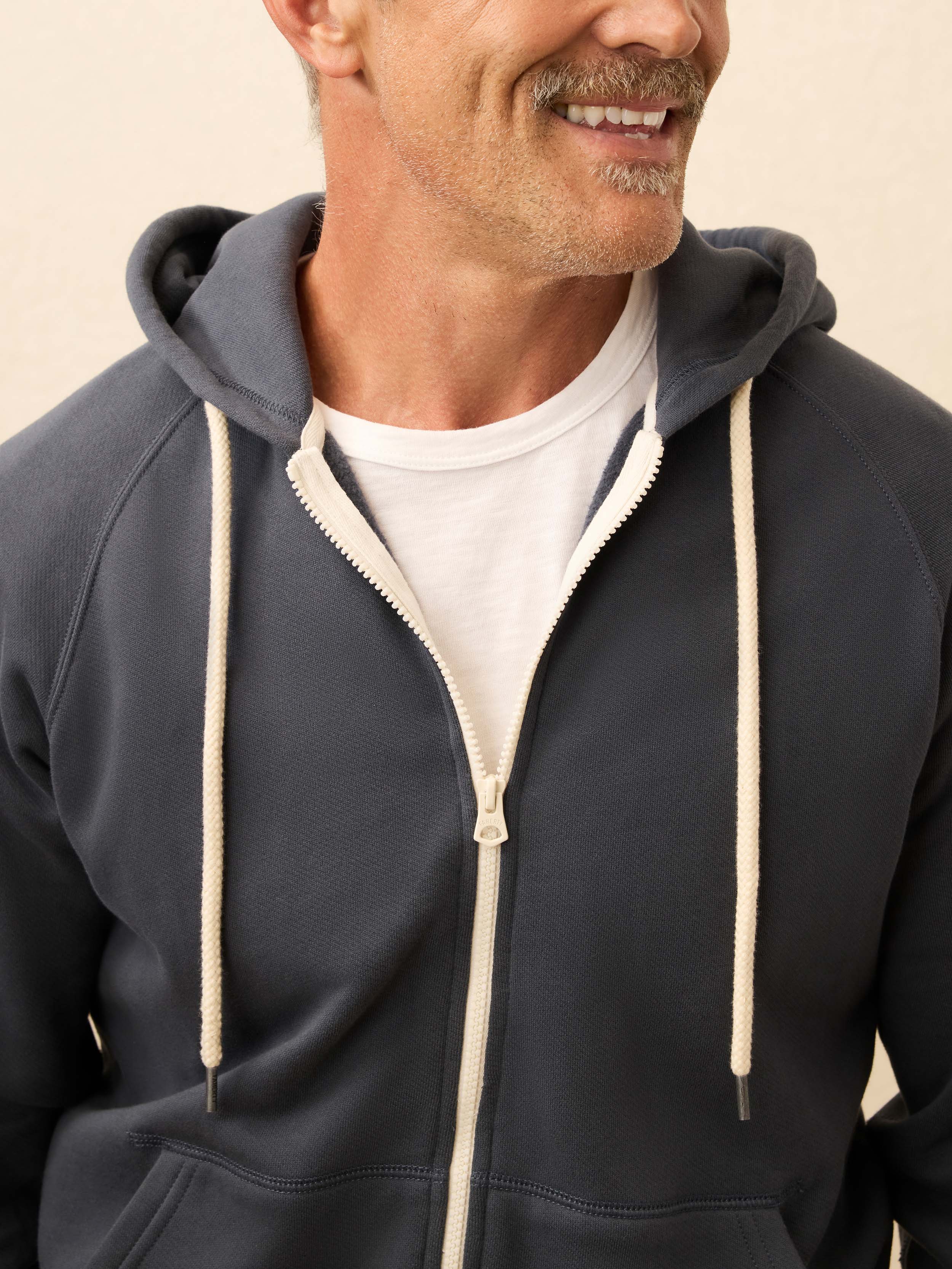Man is wearing a navy zip hoodie layered over a white shirt against a beige background