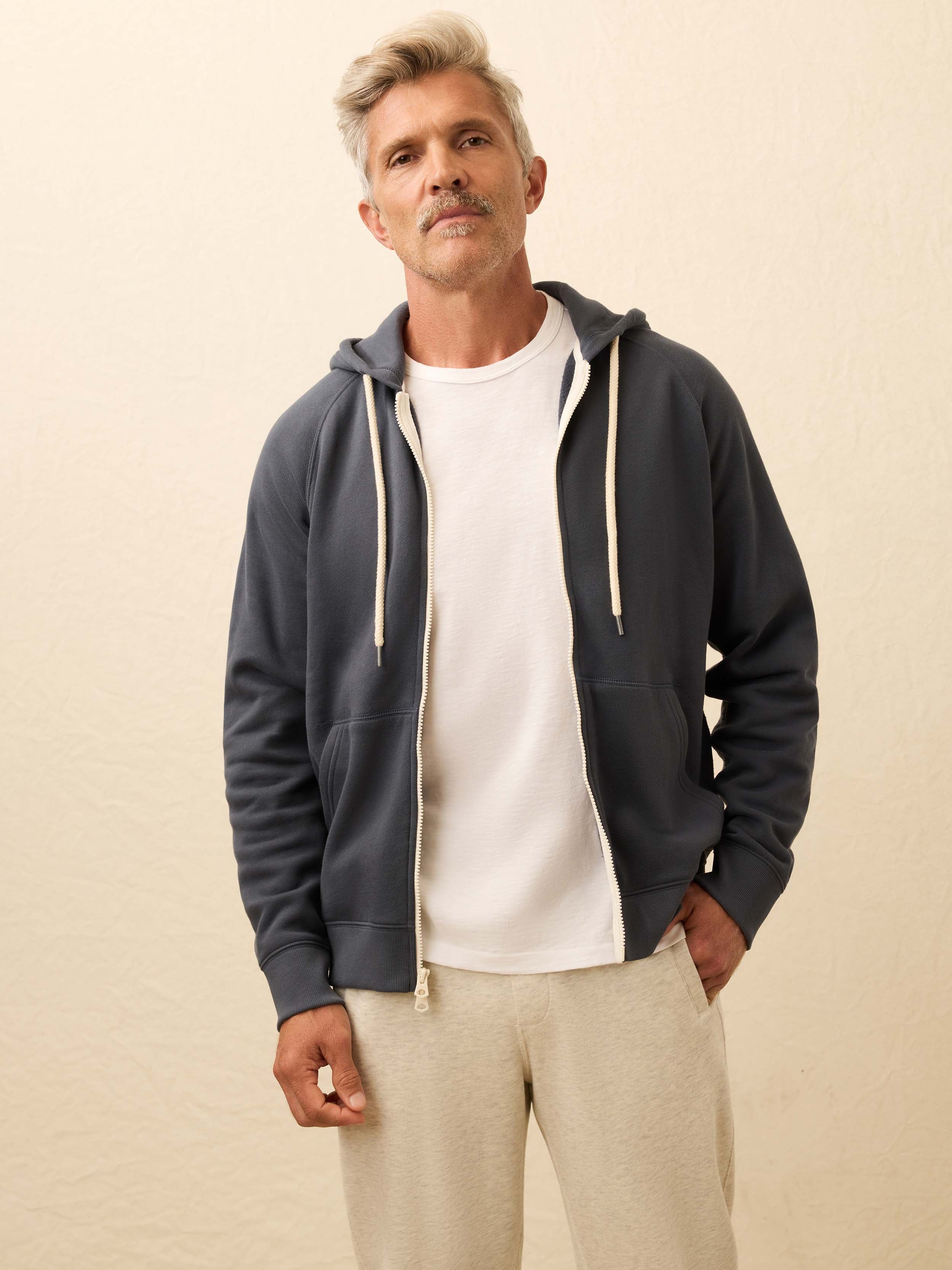 Man is wearing a navy zip hoodie layered over a white shirt against a beige background