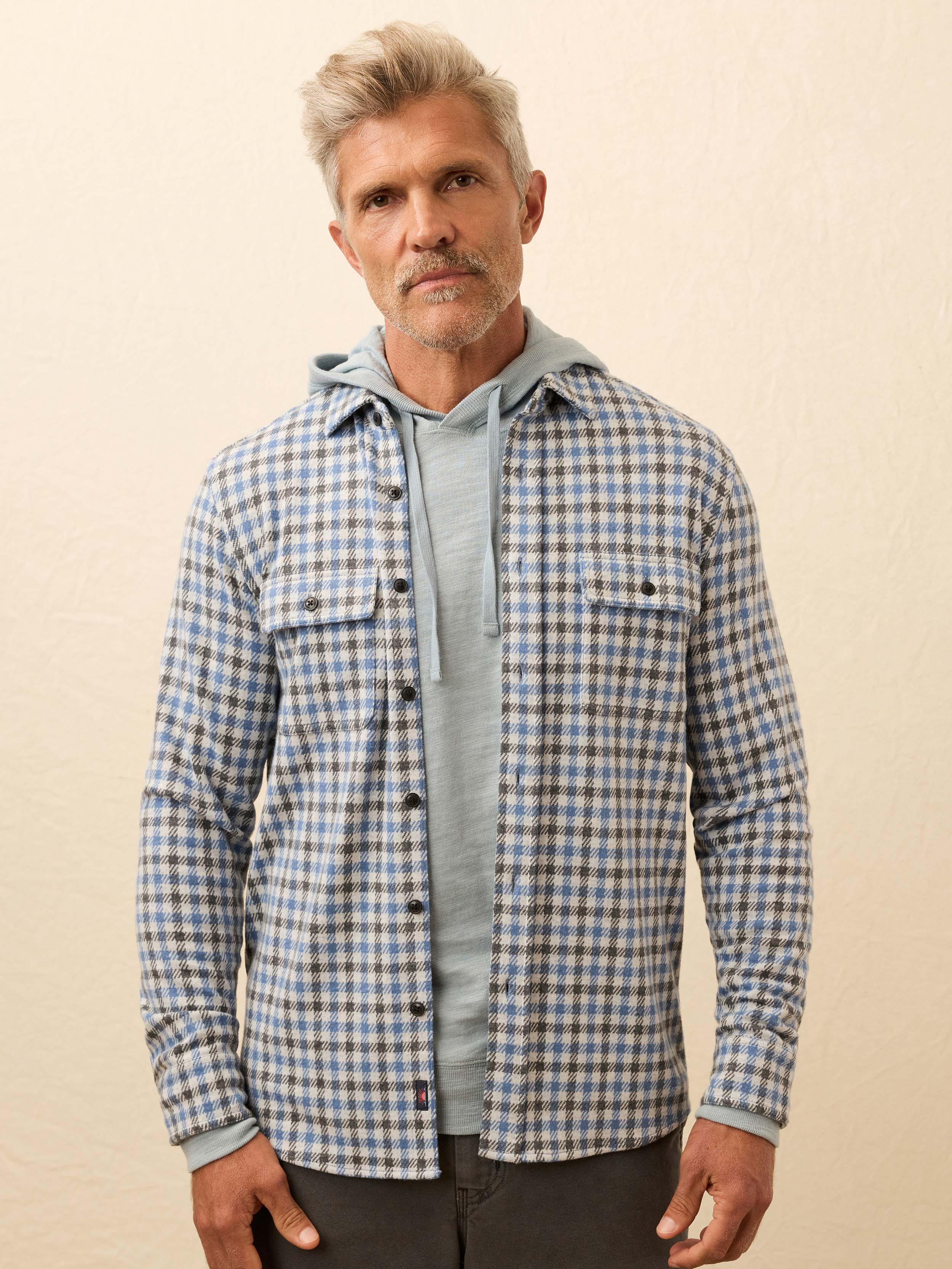 Man wearing a blue plaid shirt layered over a gray hoodie against a beige background