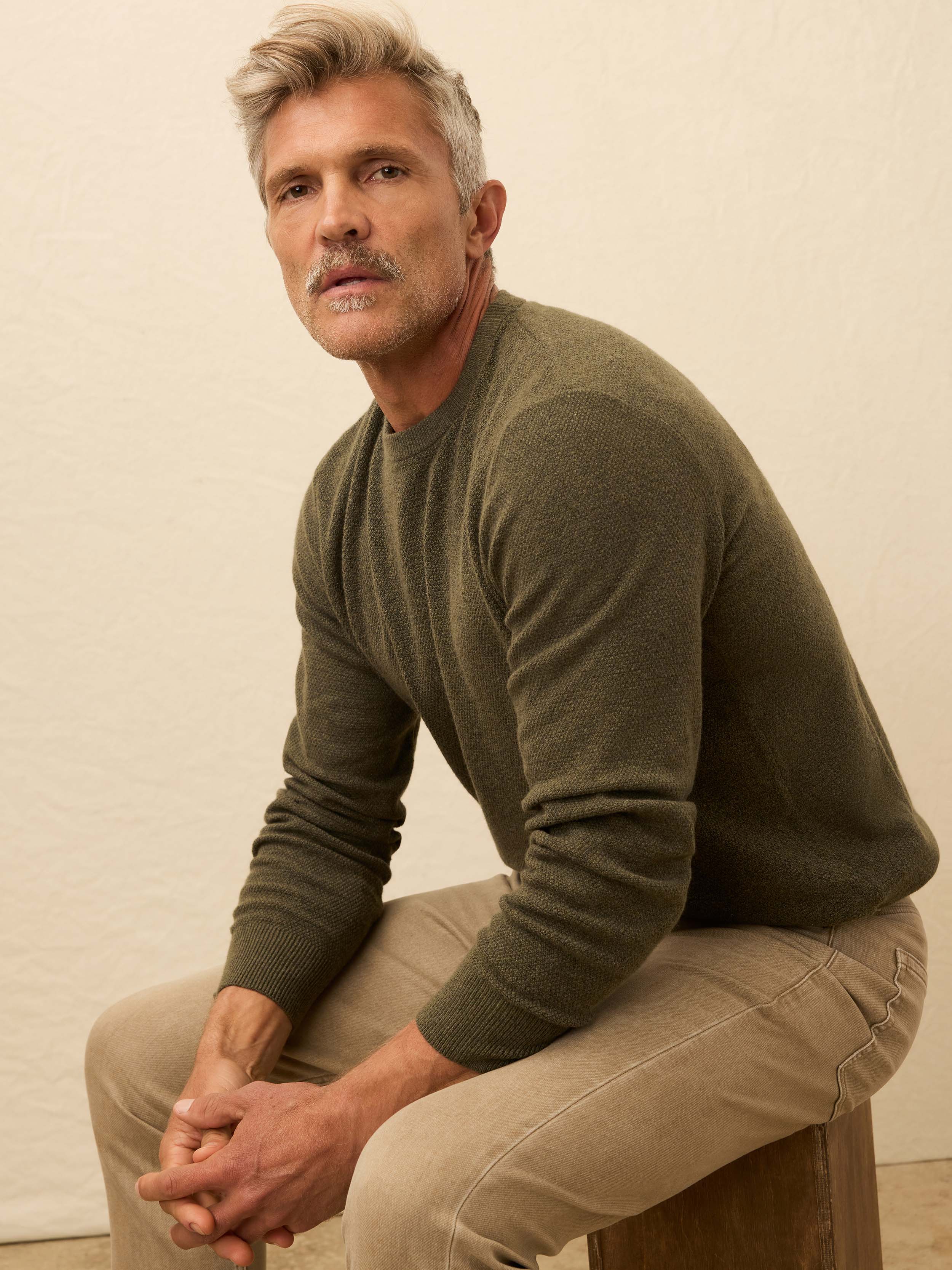 Man is wearing a green crewneck sweater with beige pants against a beige background