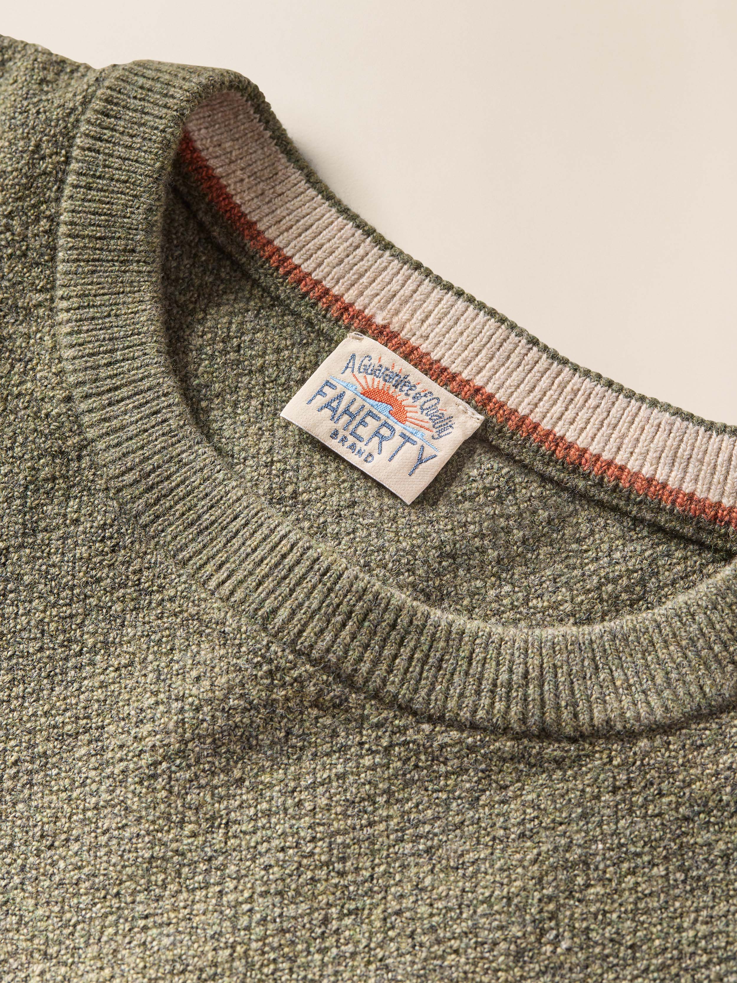 Close up of a green crewneck sweater with Faherty brand logo at the collar against a beige background