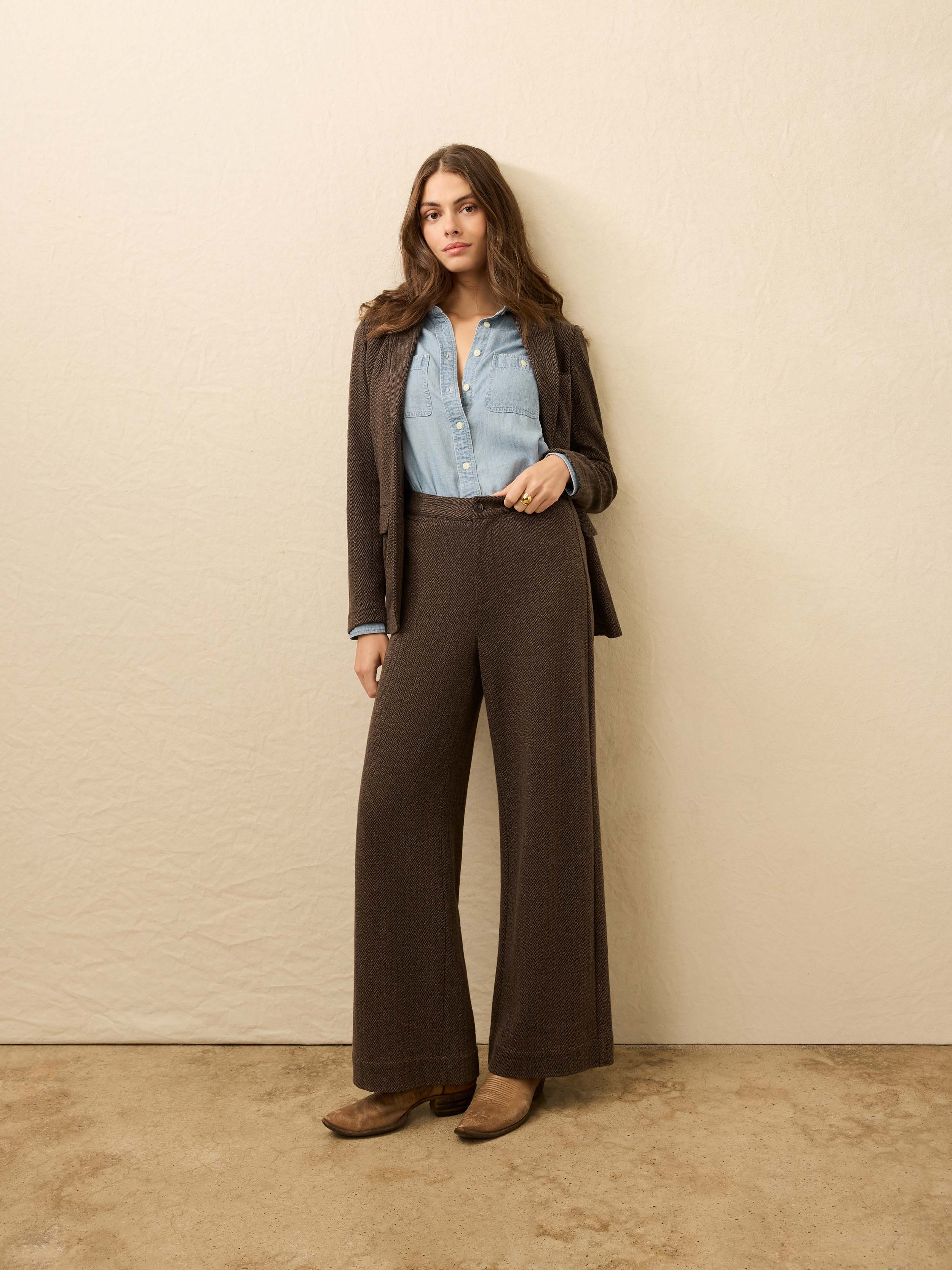 Woman is wearing brown herringbone trousers with matching blazer against a beige background