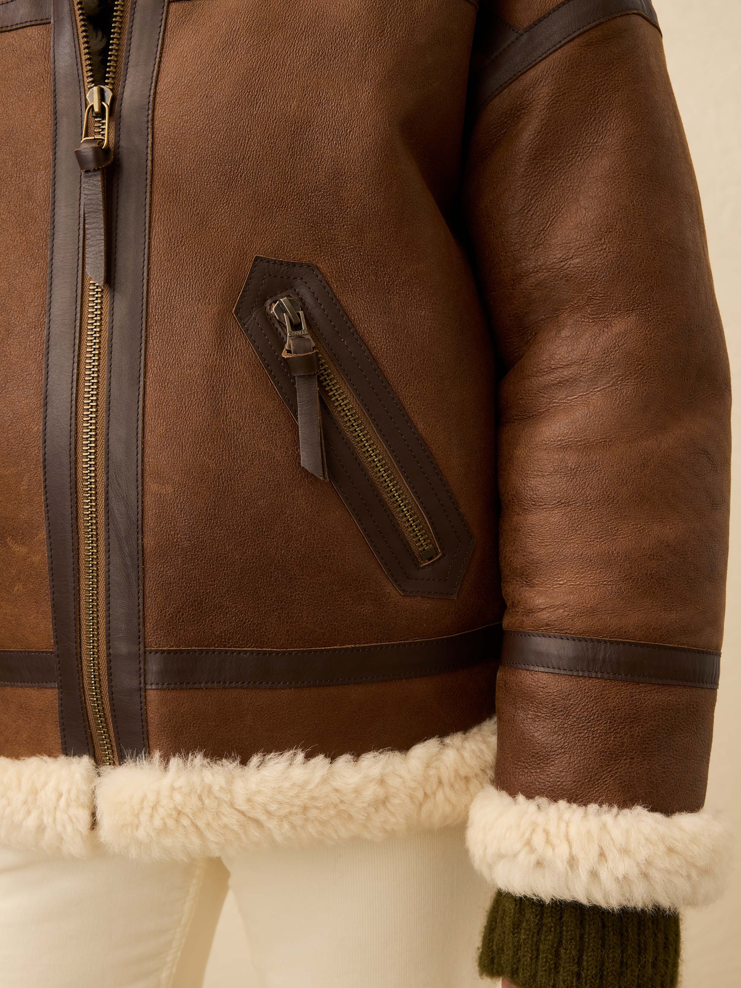 Close up of brown shearling jacket with zipper detailing with a white jean against a beige background