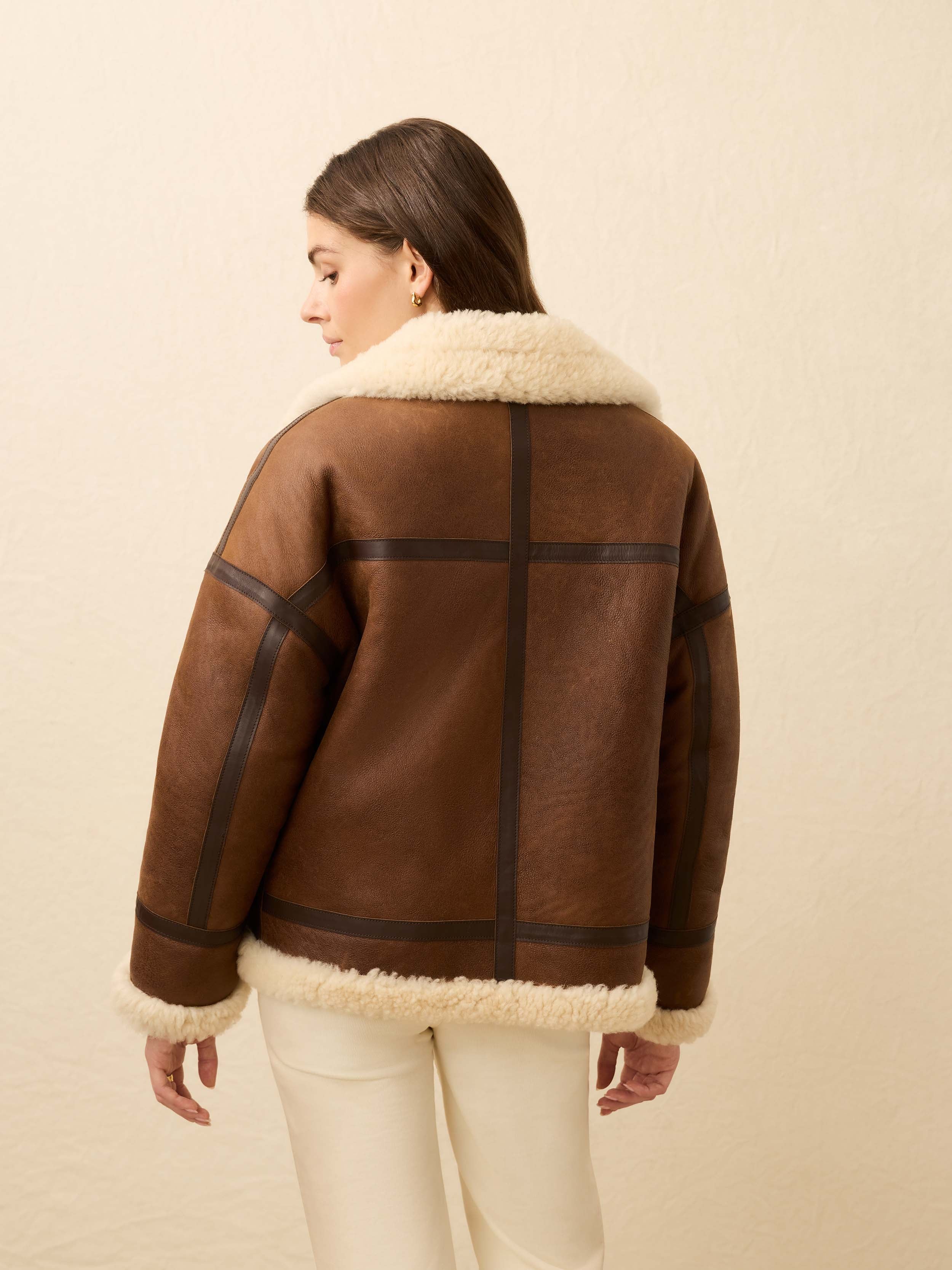 Woman is wearing a brown shearling jacket and white jeans against a beige background