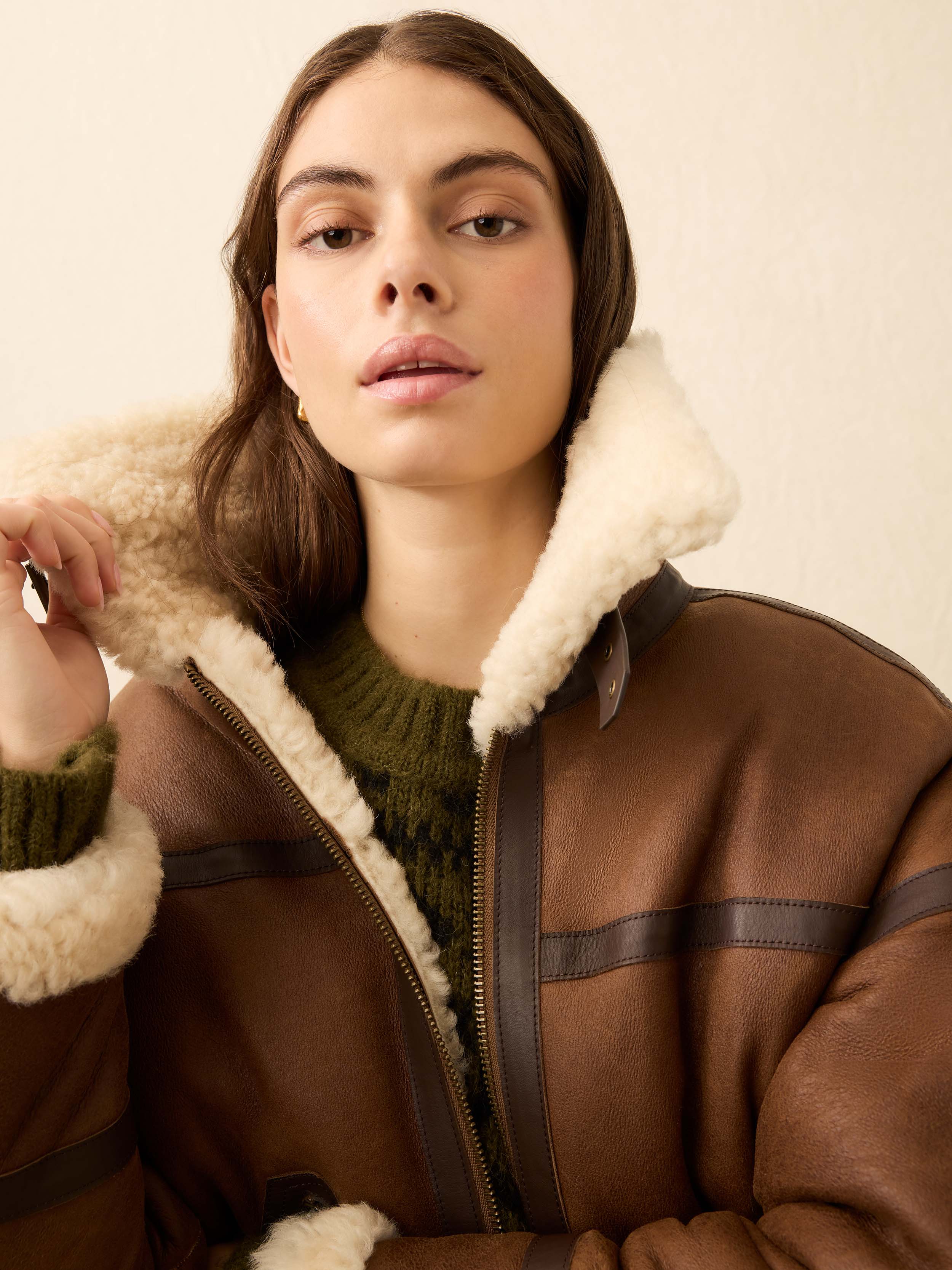 Woman is wearing a brown shearling jacket against a beige background