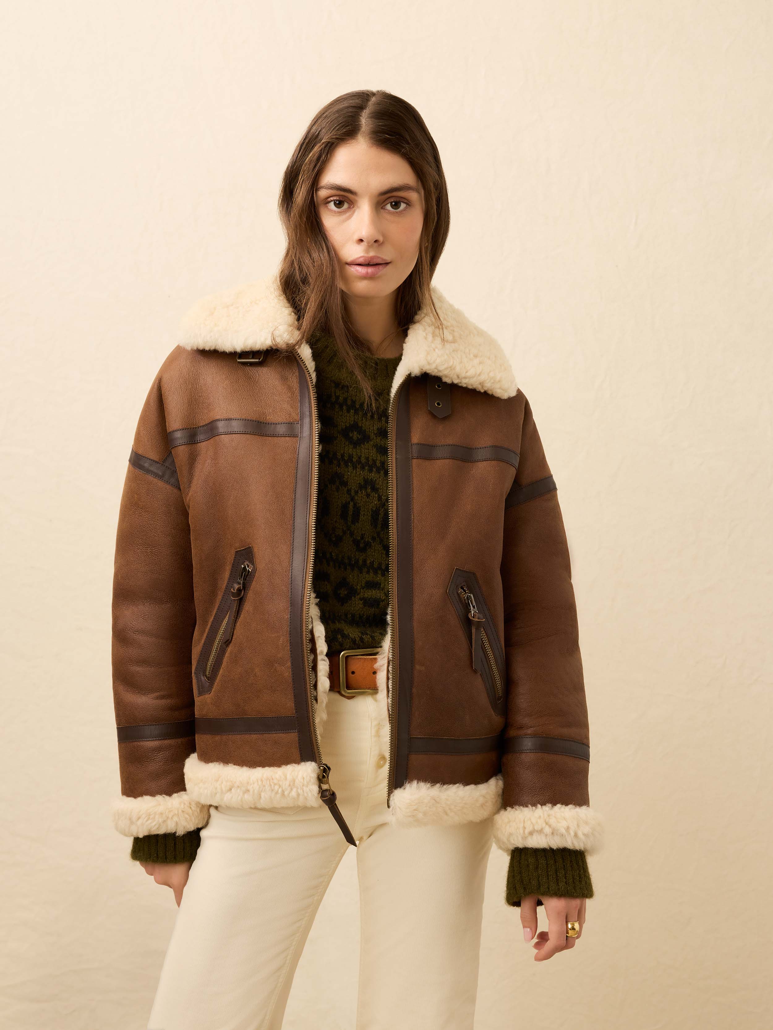Woman is wearing a brown shearling jacket and white jeans against a beige background