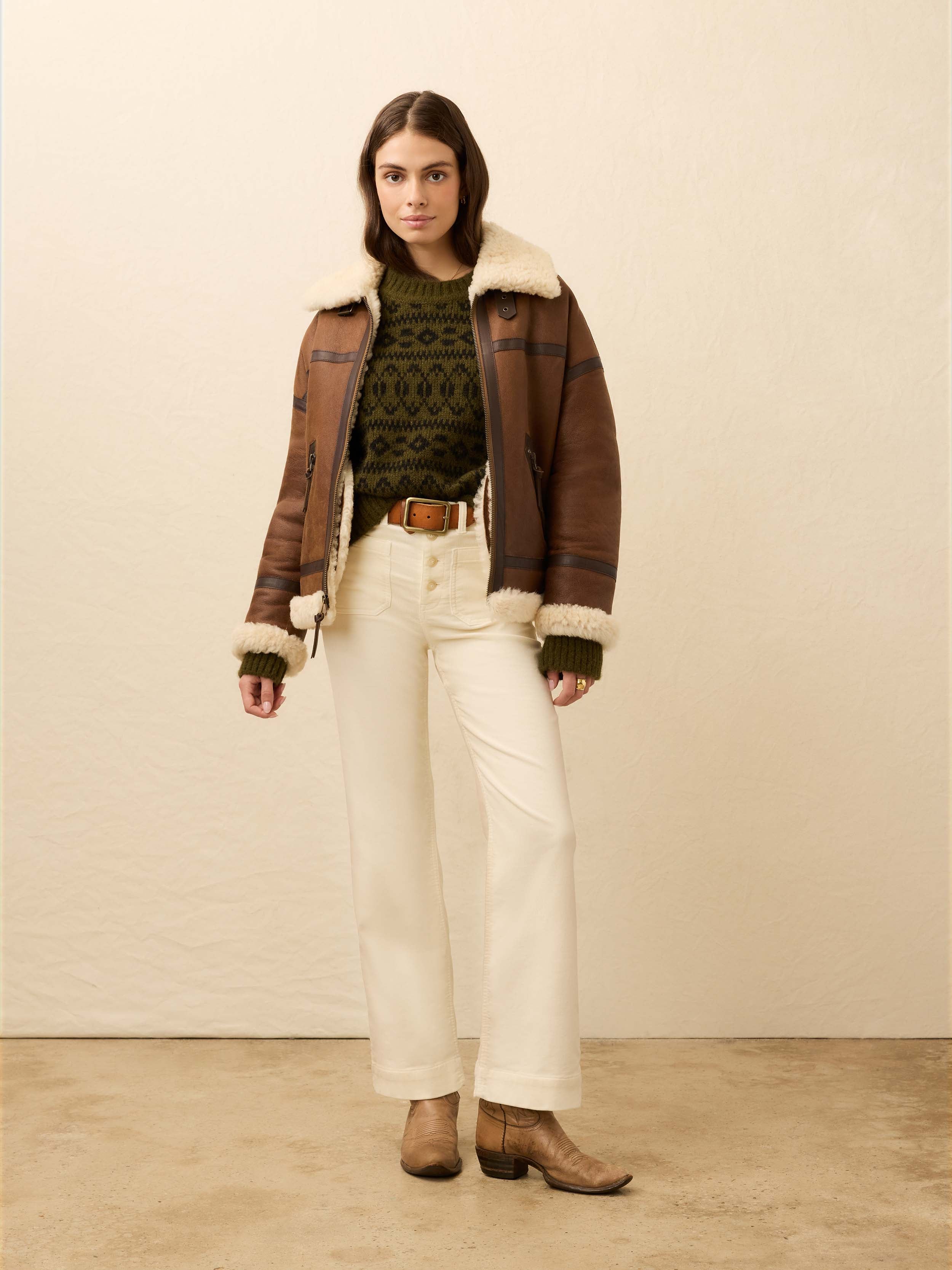 Woman is wearing a brown shearling jacket with white jeans against a beige background