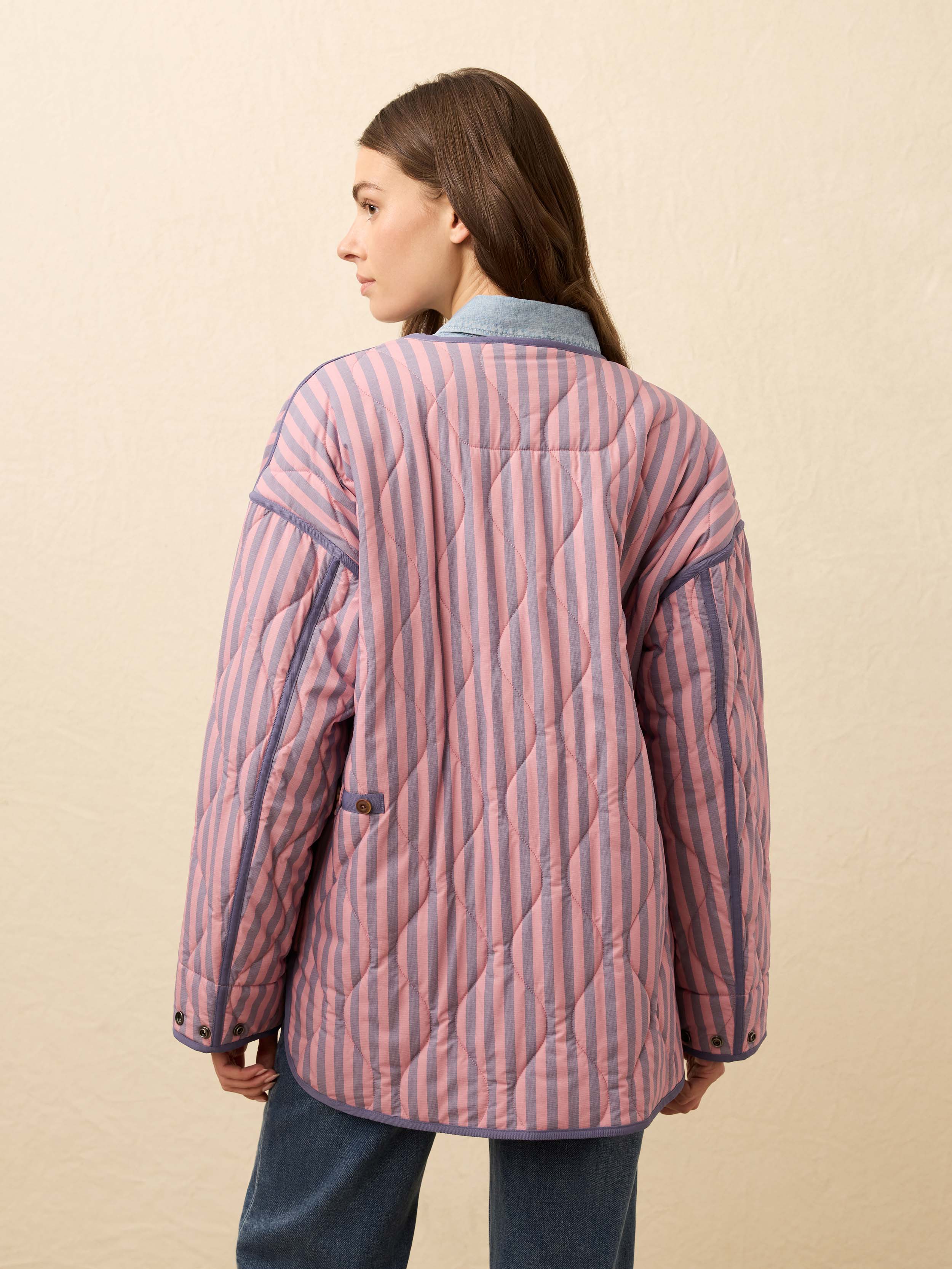 Woman is wearing a pink and blue striped quilted jacket with jeans against a beige background