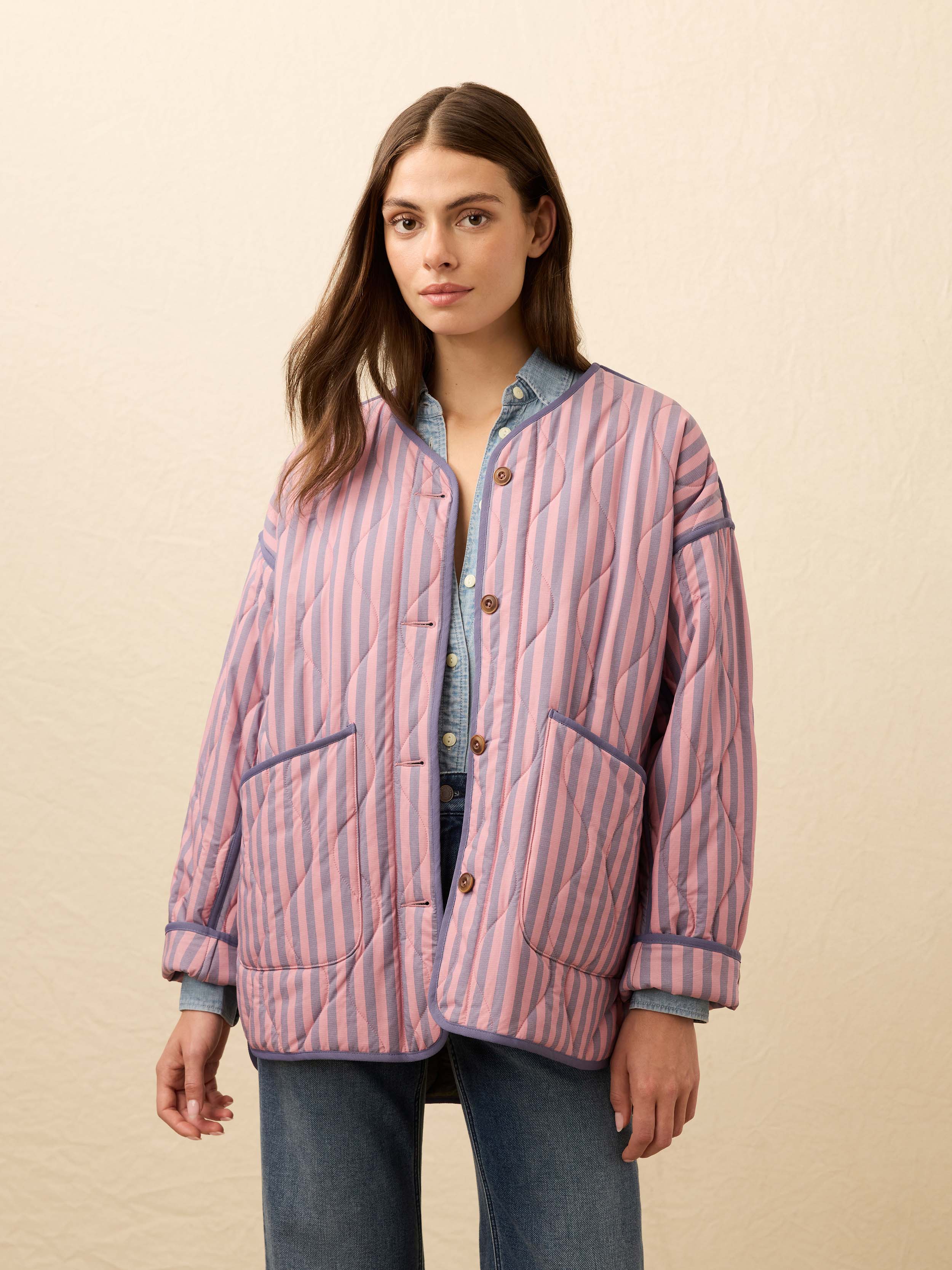 Woman is wearing a pink and blue striped quilted jacket with jeans against a beige background