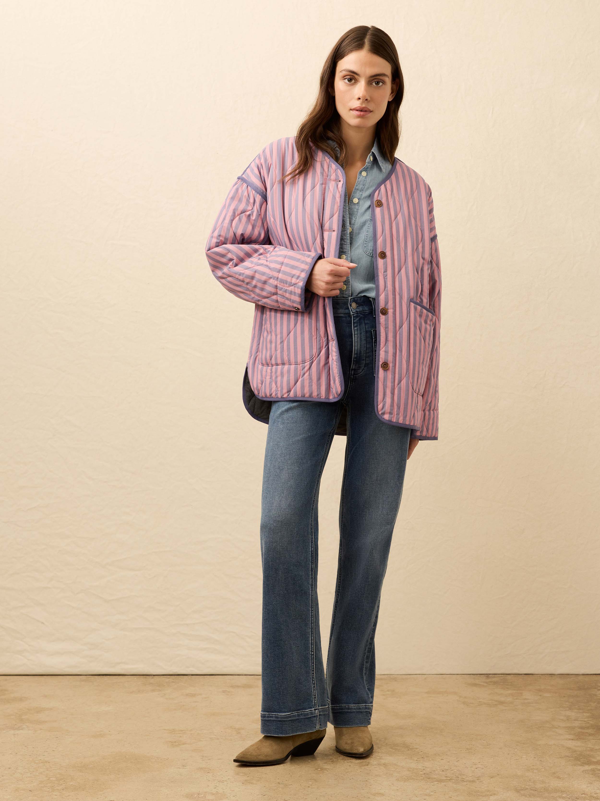 Woman is wearing a pink and blue striped quilted jacket with jeans against a beige background