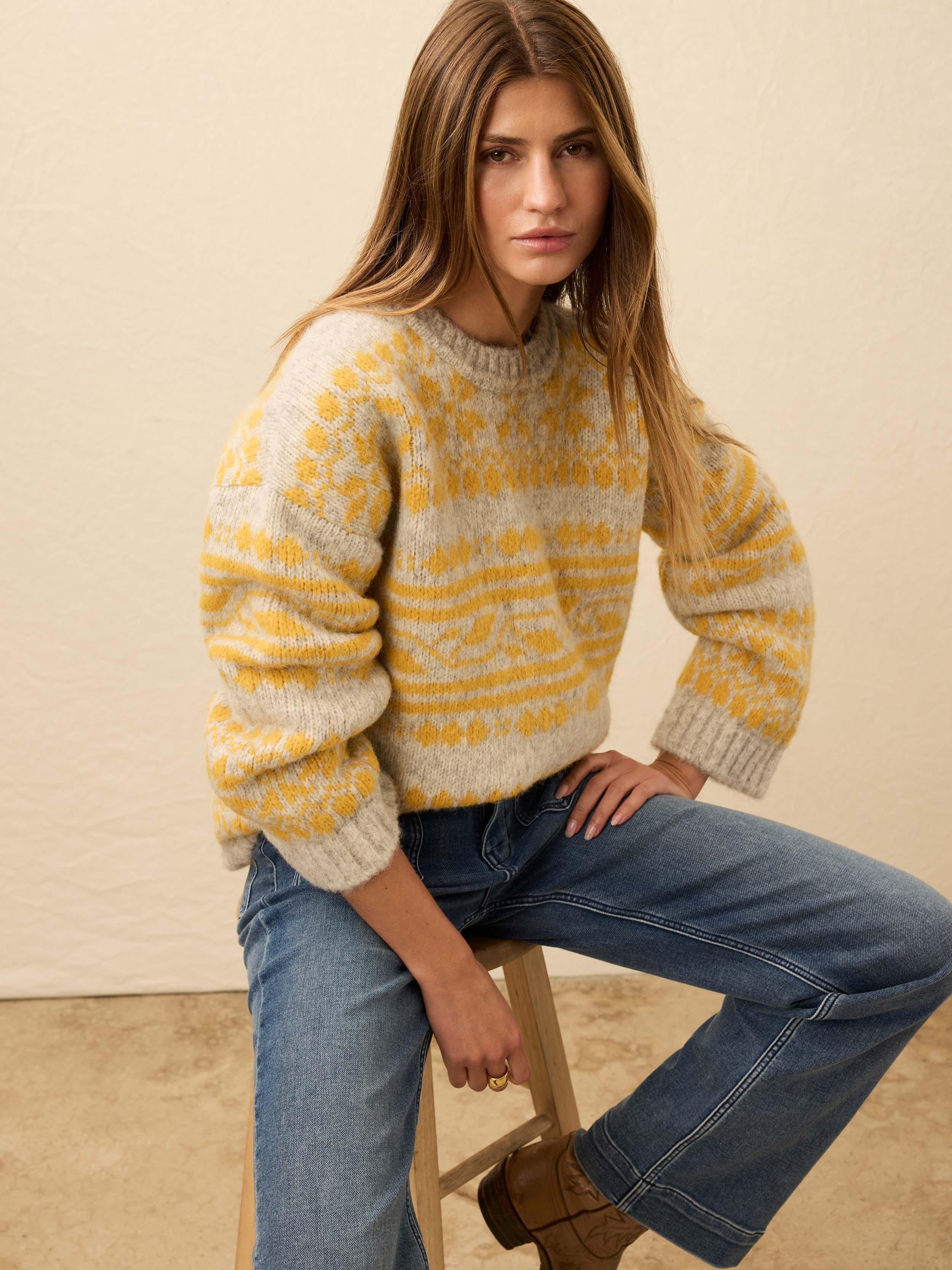 Woman is wearing a yellow fair isle sweater with blue jeans against a beige background