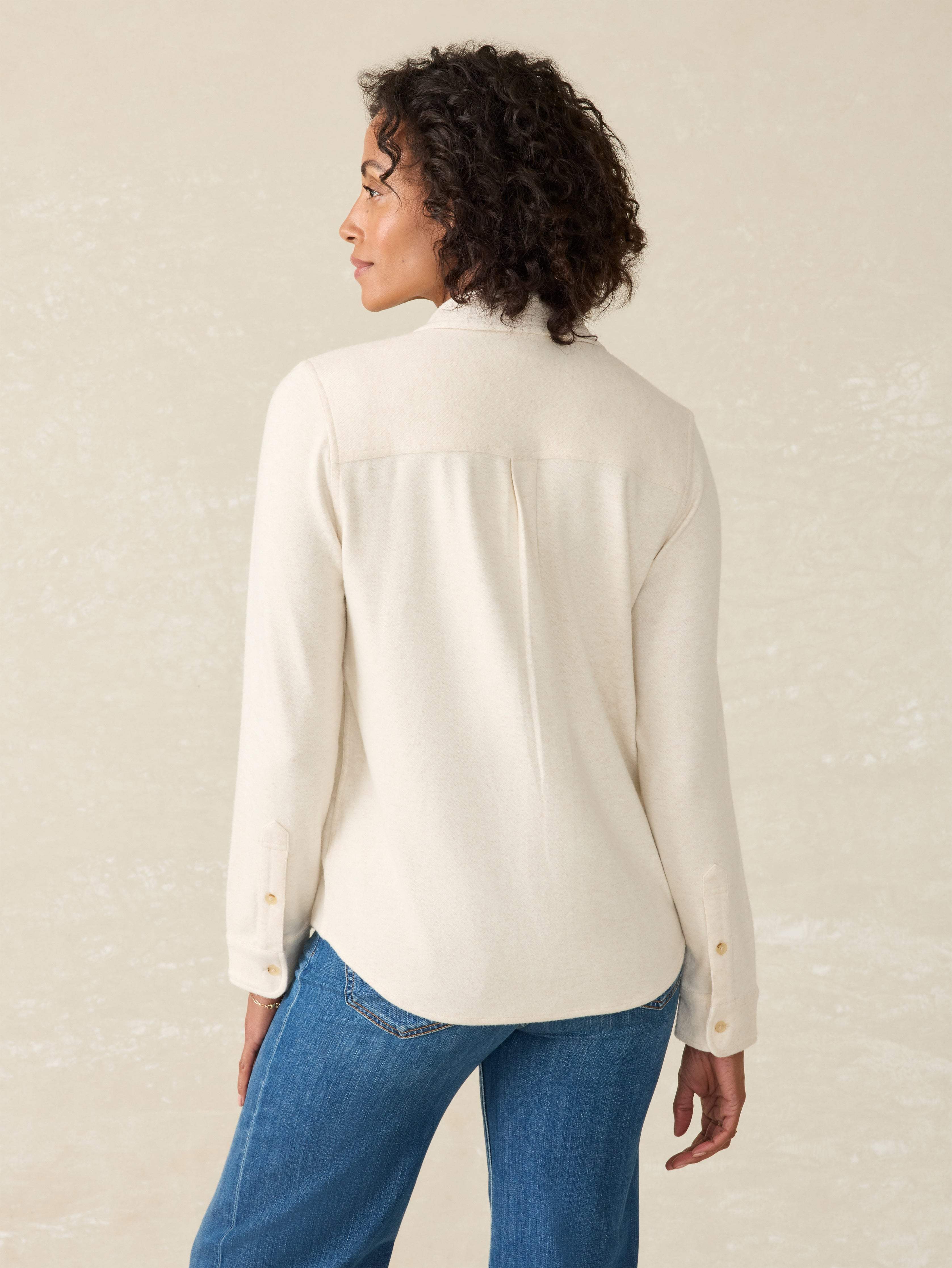 Back view of a woman wearing a cream-colored button-up shirt with Arizona State University logo on the left chest and blue jeans on a beige background