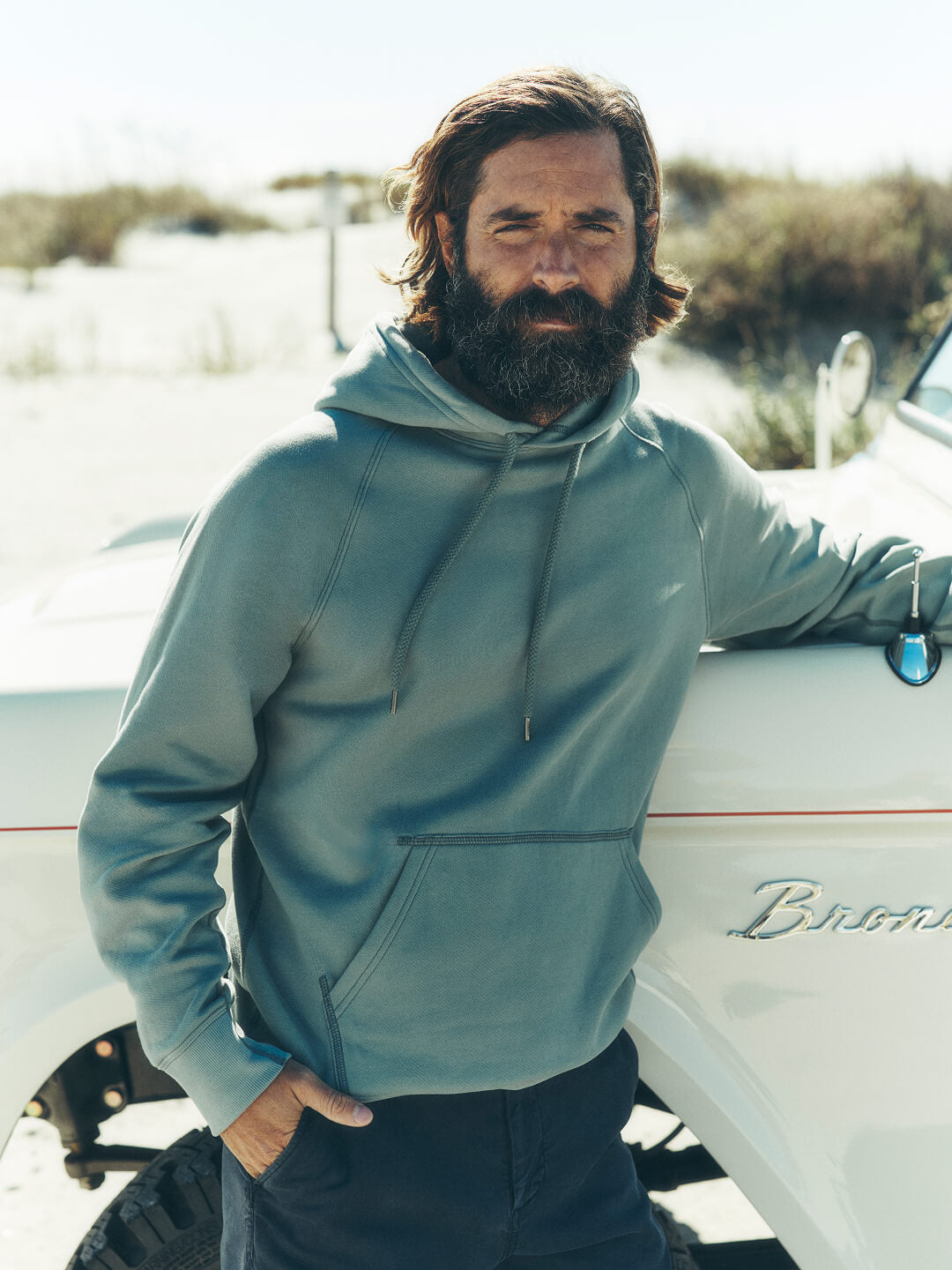 A male model leaning against a ford bronco wearing a high standard fleece hoodie