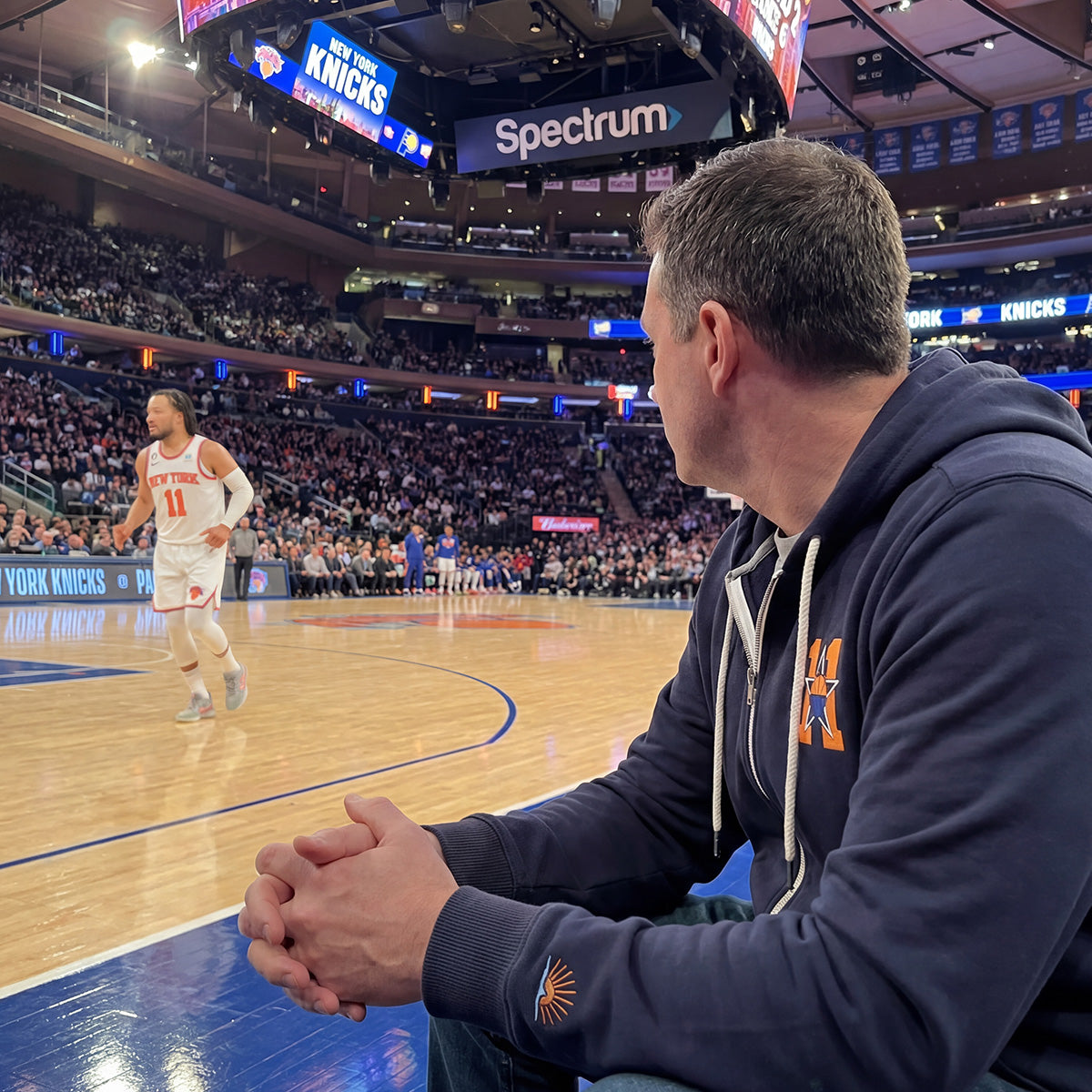 Man sitting court side at a New York Knicks game wearing a Faherty x Jalen Brunson sweatshirt while Jalen Brunson is playing basketball in the background.