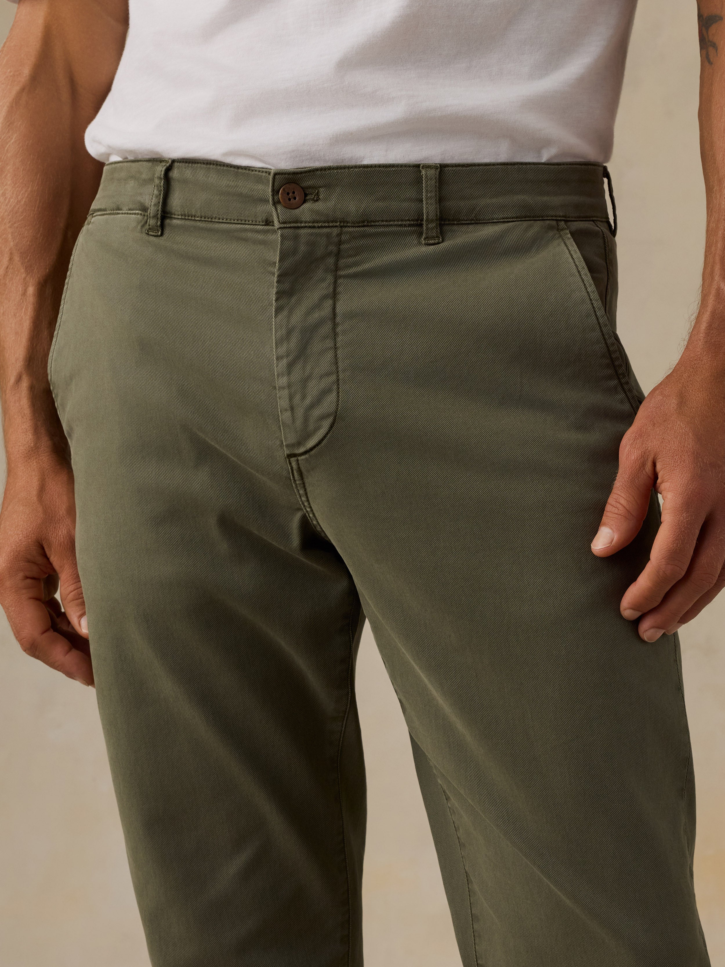 A close-up of a man wearing olive green chinos with a white t-shirt, showing the front waist, belt loops, button, and front pockets.