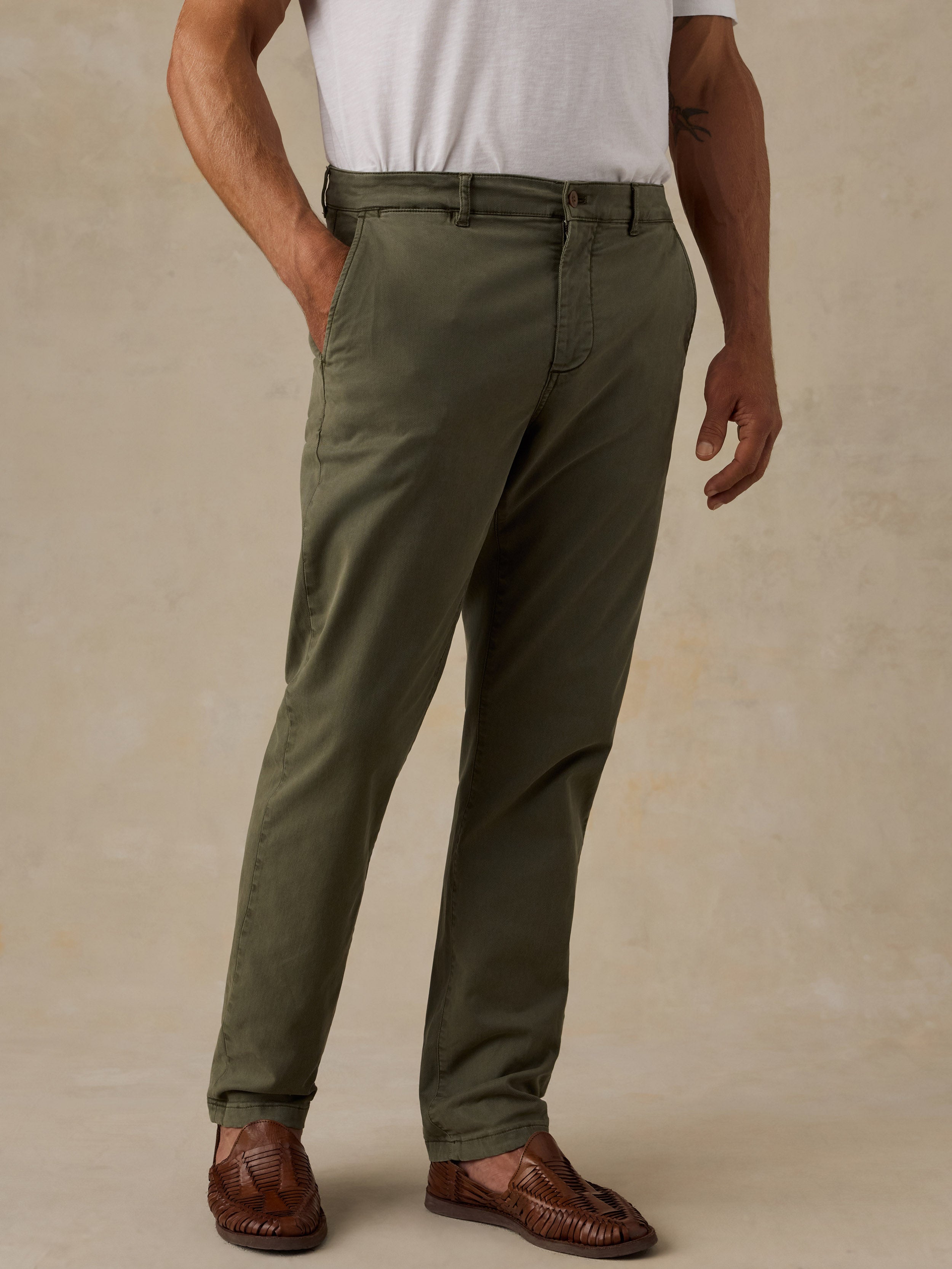 A man wearing olive green slim-fit trousers, a white t-shirt, and brown leather loafers, standing against a textured beige studio background.