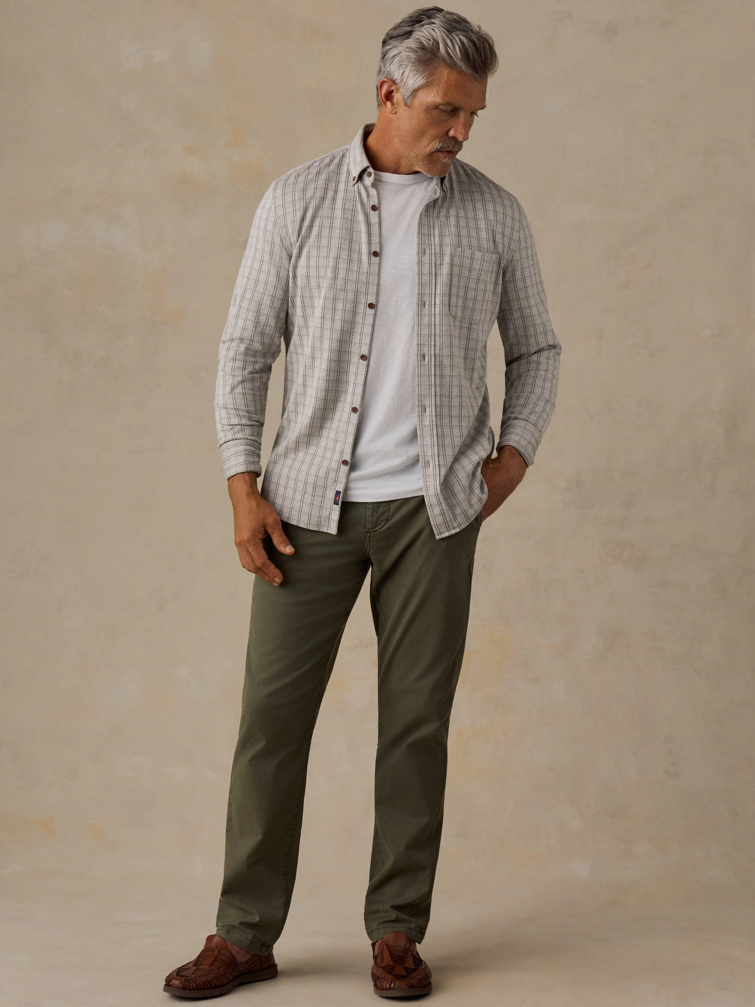 A man wearing a light gray and white checkered button-down shirt over a white t-shirt, paired with olive green pants and brown shoes, standing against a textured beige studio backdrop.