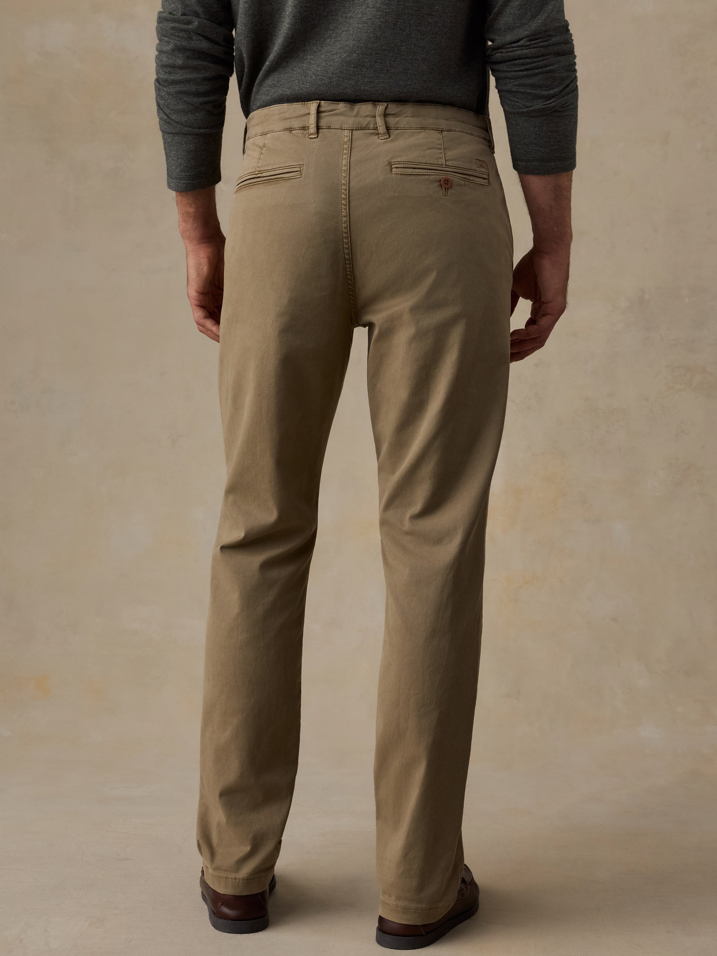 A man viewed from the back wearing straight-leg khaki trousers, a dark gray long-sleeve shirt, and brown shoes, standing against a textured beige studio backdrop.