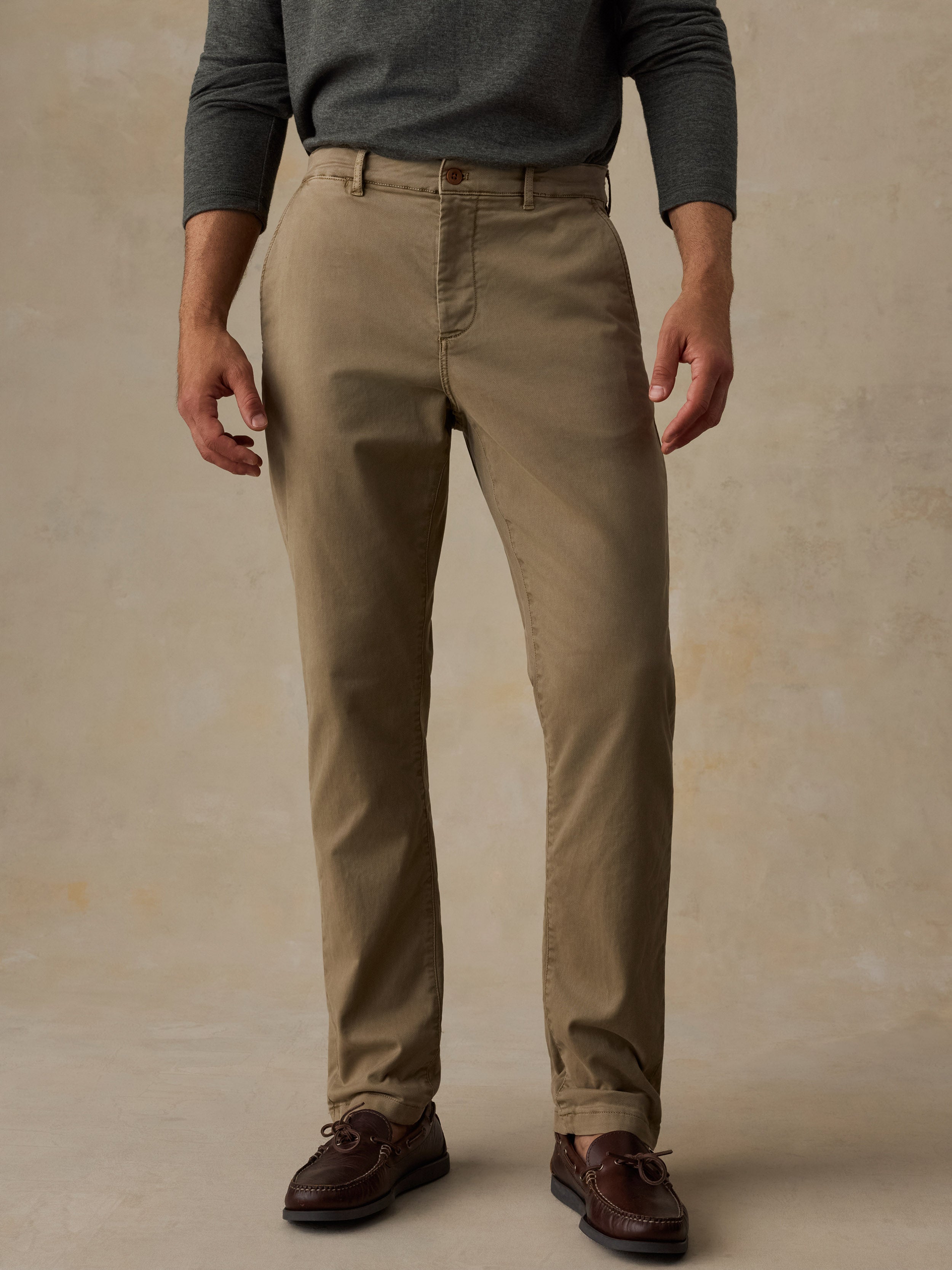 A man wearing khaki chino pants, a dark gray long-sleeve shirt, and brown leather shoes, standing against a textured beige studio backdrop.