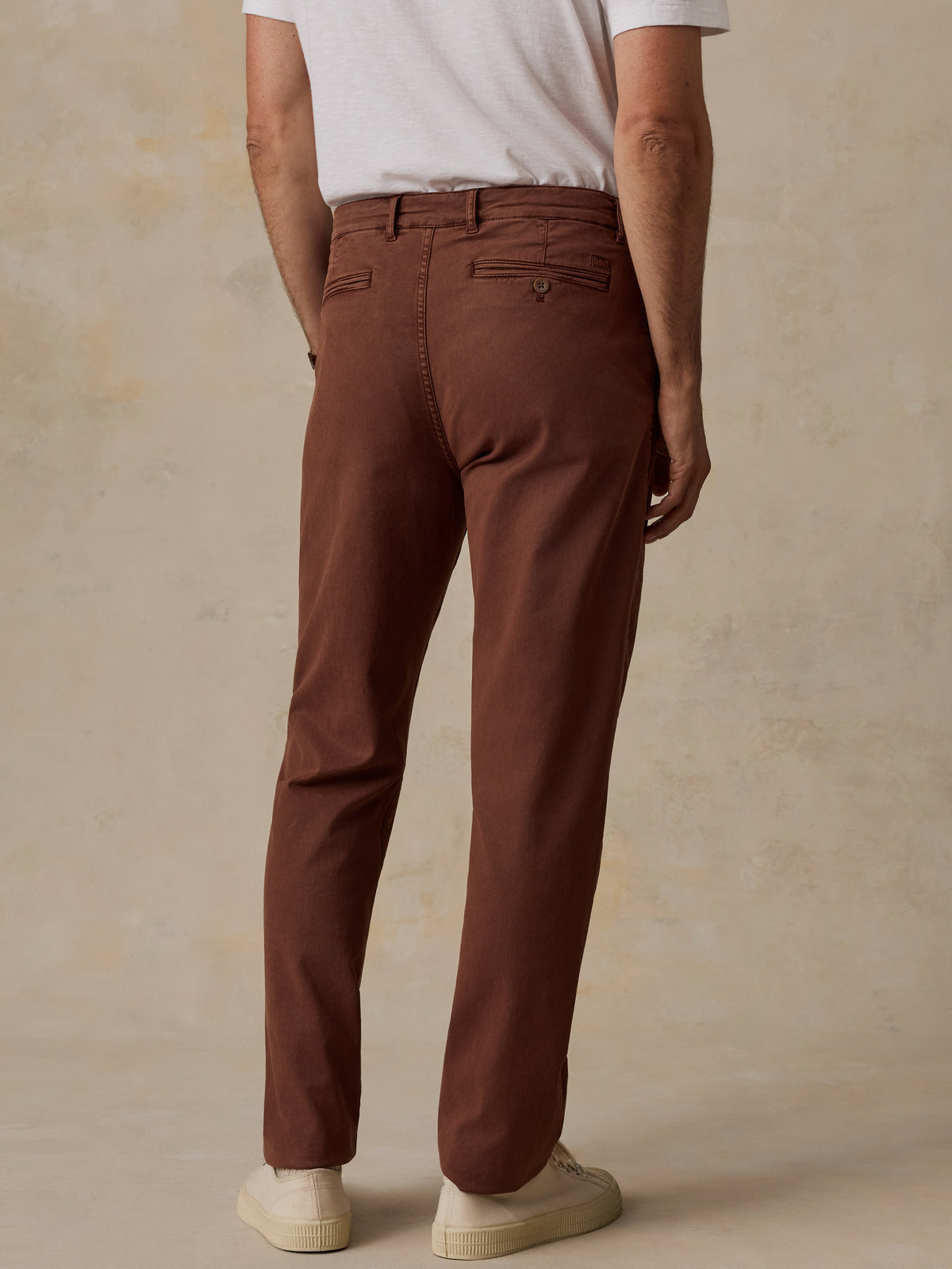 A person shown from the back wearing brown straight-leg chino pants, a white t-shirt, and off-white sneakers, standing against a textured beige studio backdrop., image 4 of 7