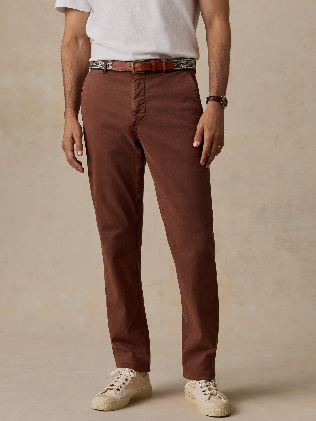 A man wearing slim-fit brown chinos with a brown leather belt, a white t-shirt, and cream sneakers, standing against a textured beige studio backdrop., image 2 of 7