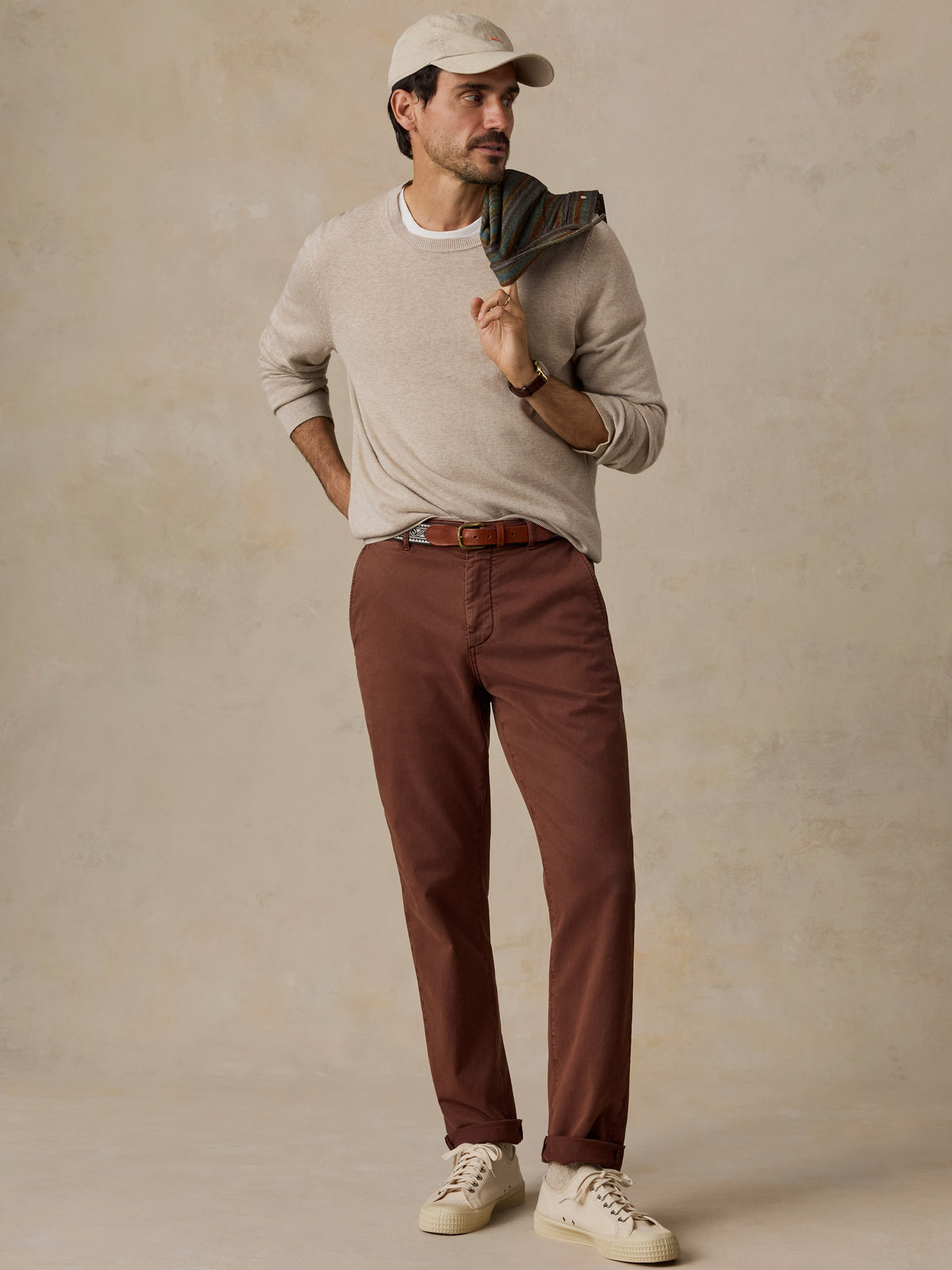 A man modeling a light beige crewneck sweater, rust-brown pants, a beige baseball cap, and cream sneakers, standing against a textured beige studio backdrop., image 7 of 7