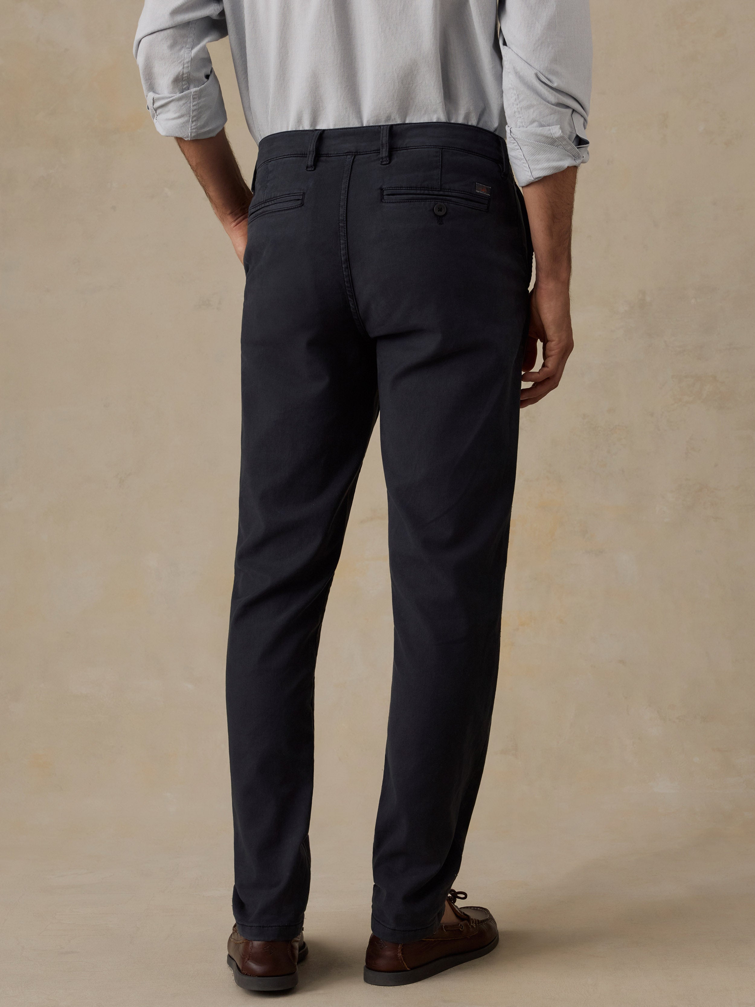 Rear view of a man wearing black trousers, a light gray button-down shirt, and brown leather loafers, standing against a textured beige studio backdrop.
