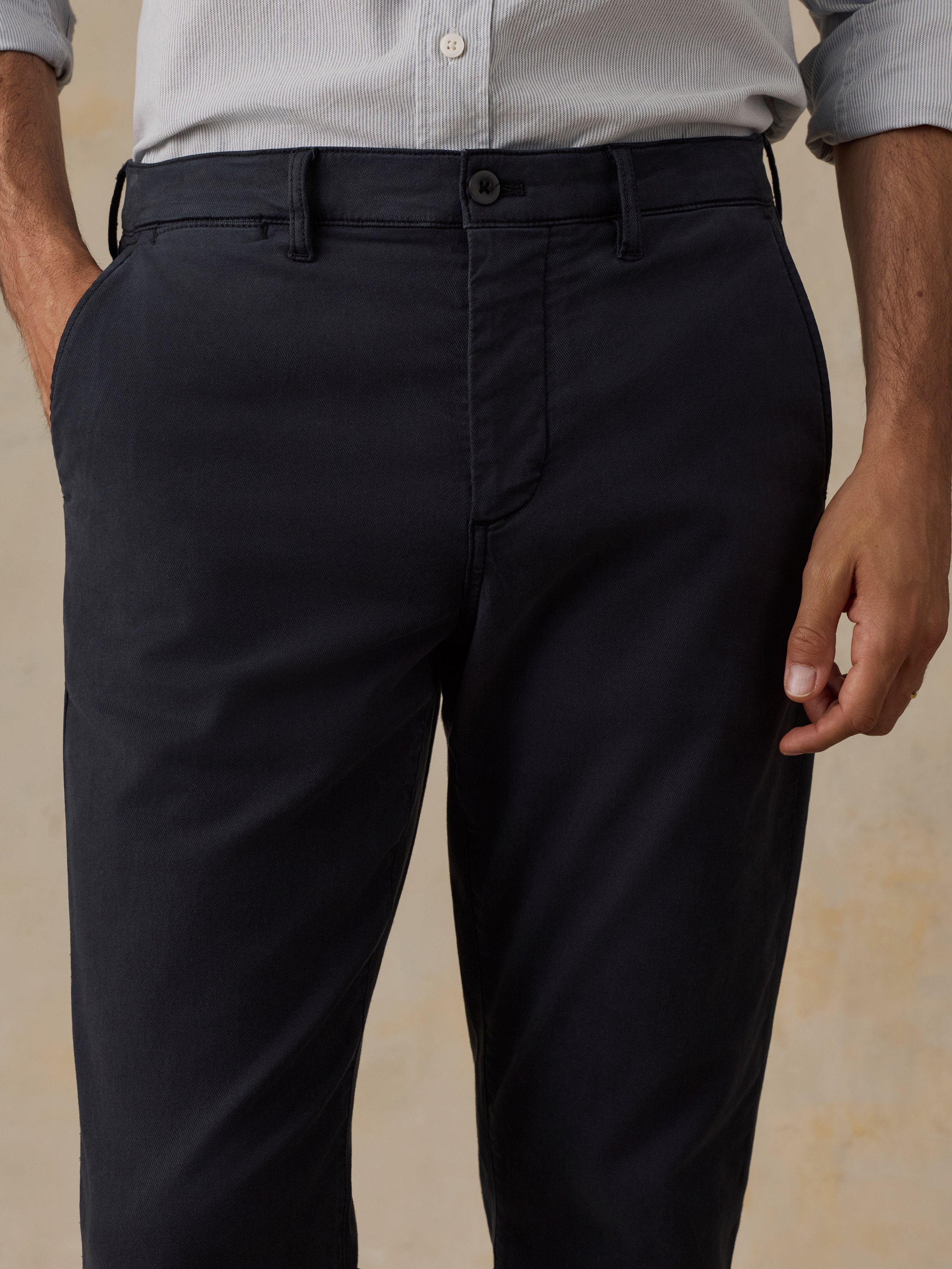 A close-up of a man wearing dark charcoal gray or black chino pants with a light gray button-down shirt, shown from the waist down against a warm beige background.