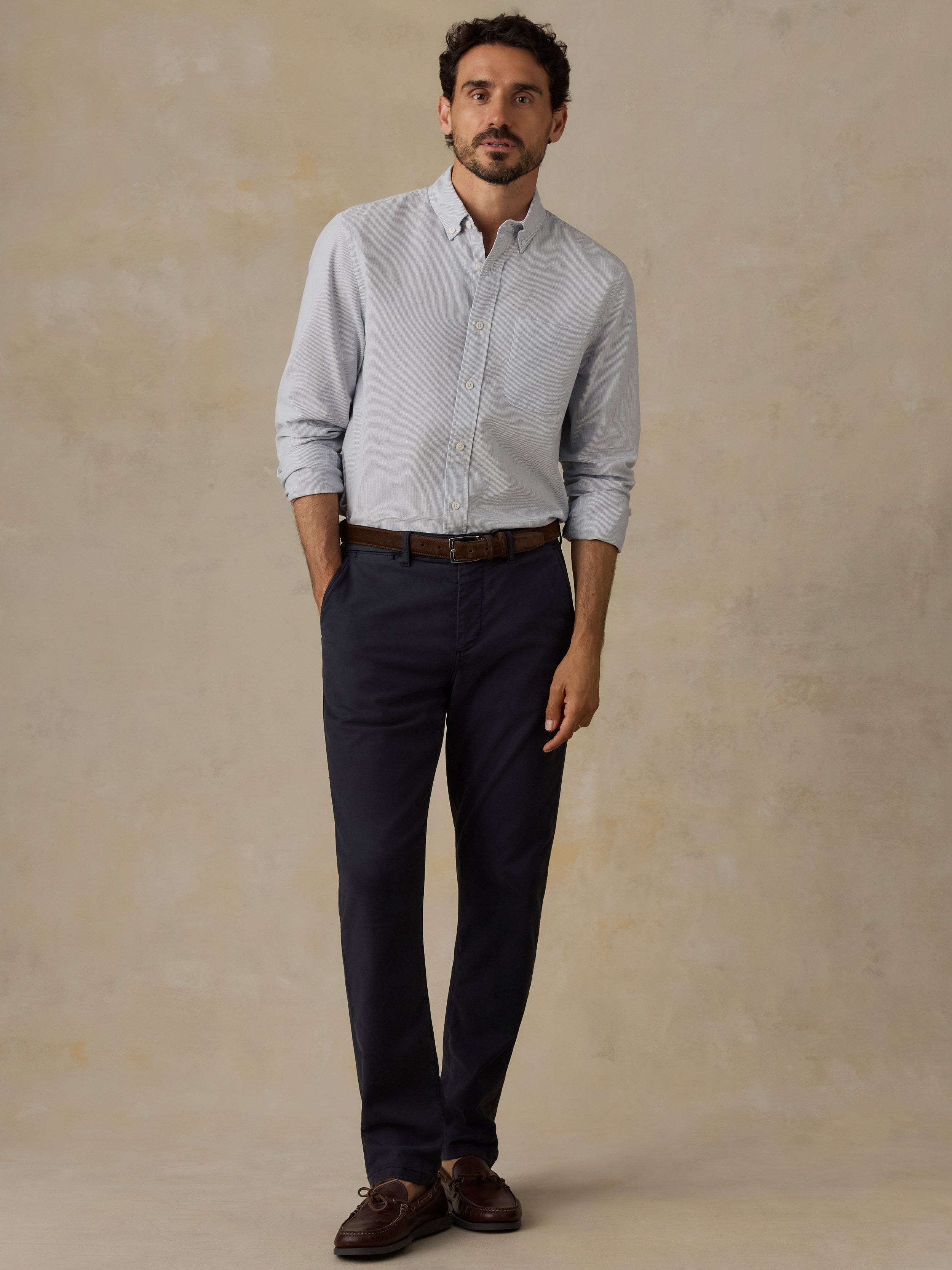 A man wearing a light blue long-sleeve button-down shirt, black trousers, a brown belt, and brown leather shoes, standing against a textured beige studio backdrop.